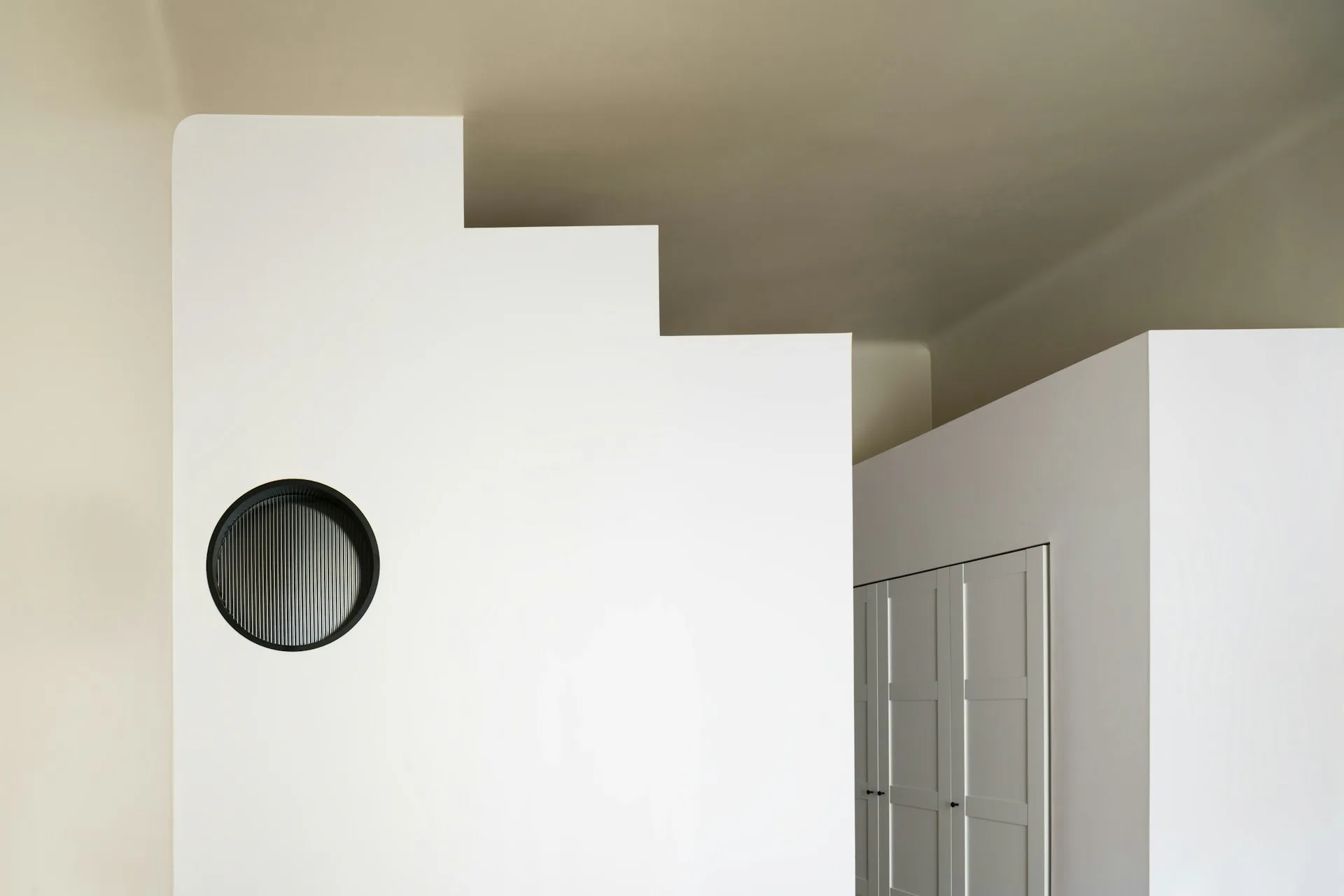 Minimalist interior with stepped white walls, a circular dark vent, and metallic door frame, featuring synthetic lighting and clean geometric forms by Alepreda Architecture.