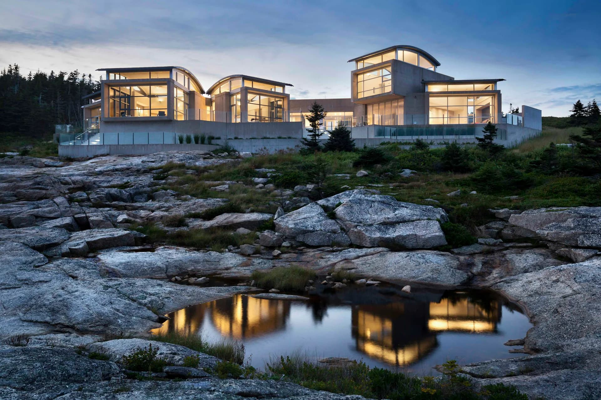 Modern residential complex with illuminated curved glass facades nestled among rocky terrain and evergreens at dusk, reflecting in a natural pool by Alexander Gorlin Architects.