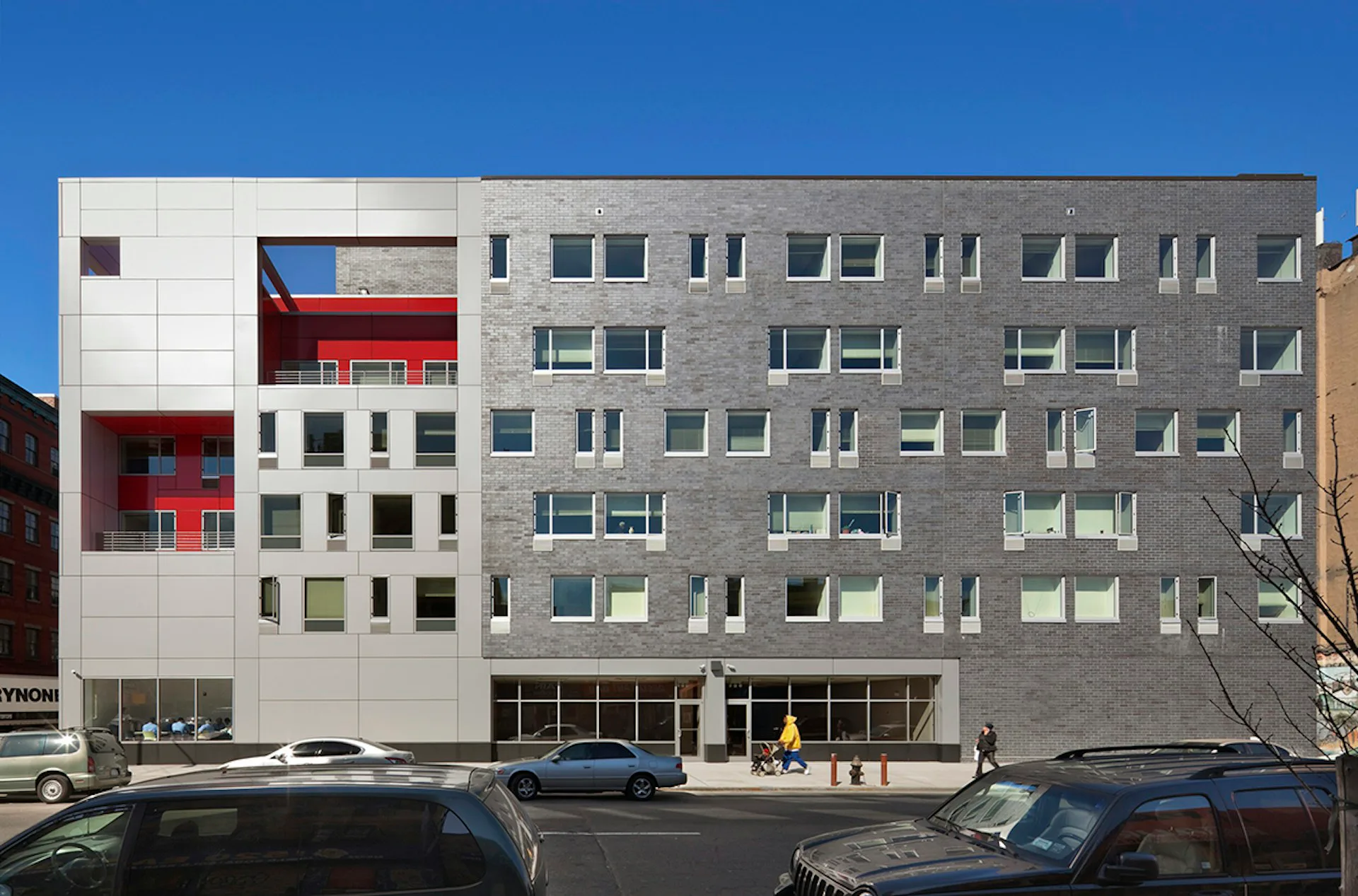 Modern multi-story mixed-use building facade with white and gray panels, red interior details visible through windows, ground-floor retail spaces, and blue sky backdrop by Alexander Gorlin Architects.