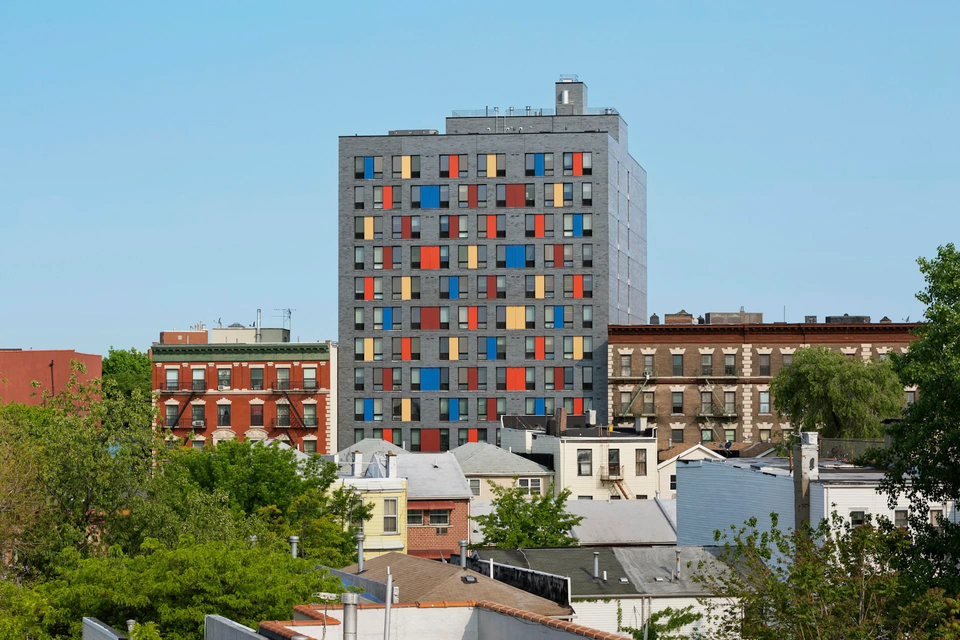 Colorful residential tower with multicolored window panels rises above lower-rise brick buildings and green trees in urban streetscape by Alexander Gorlin Architects.