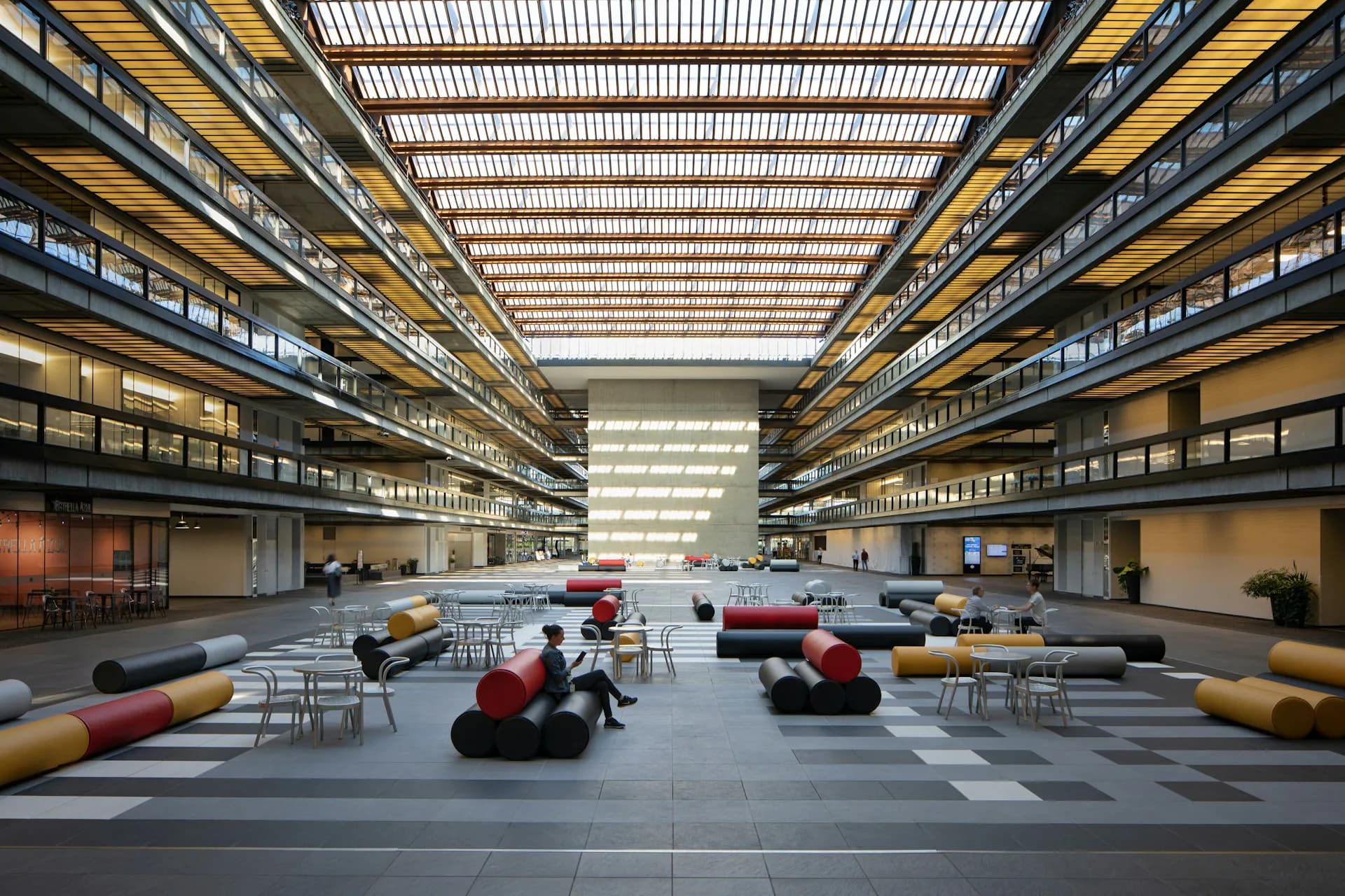 A bright industrial warehouse interior with yellow overhead cranes, multi-level storage racks, and colorful cylindrical objects arranged on a gray floor, designed by Alexander Gorlin Architects.