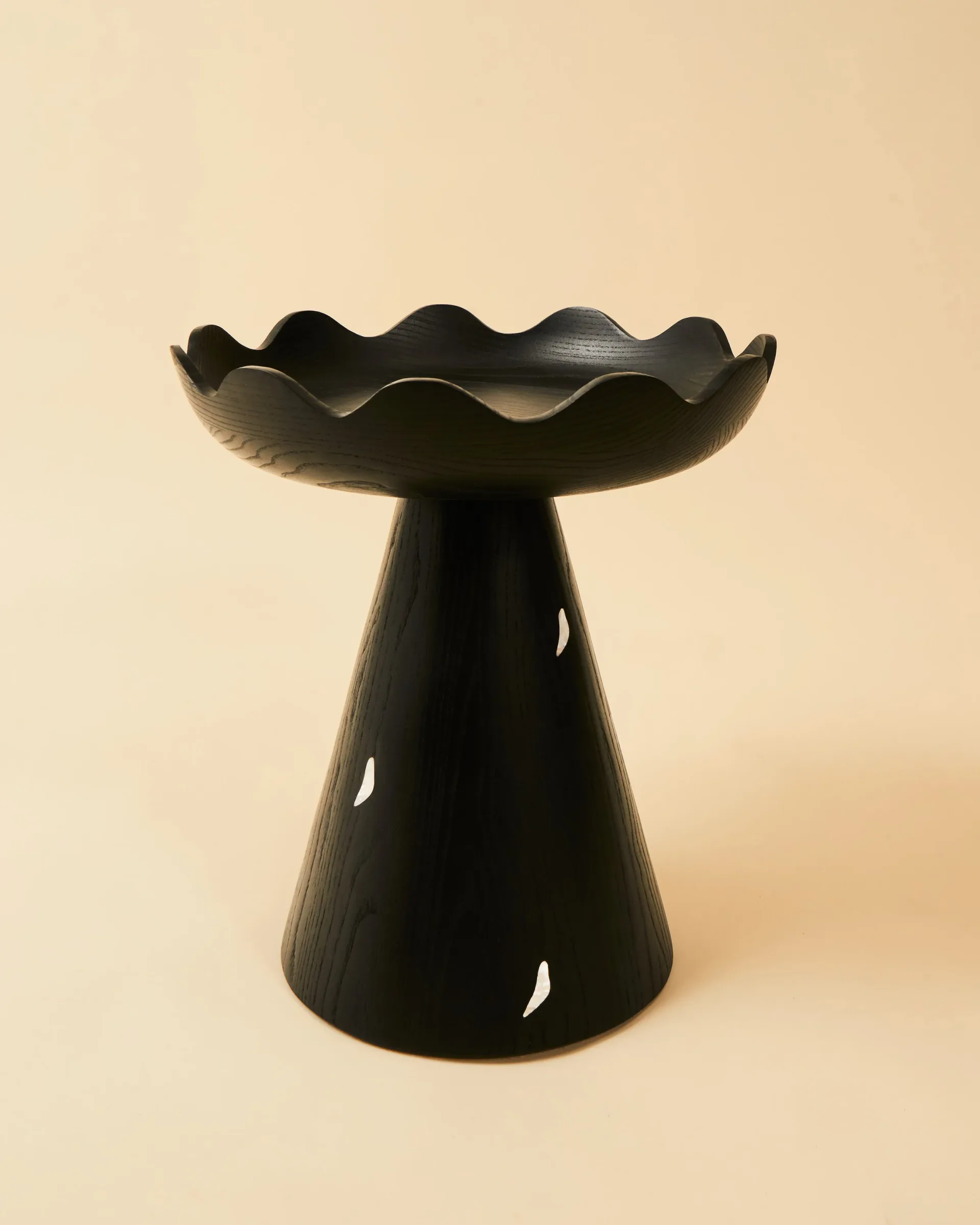 Black sculptural pedestal bowl with wavy rim and decorative cutouts, photographed against a neutral cream background by Alexis & Ginger.