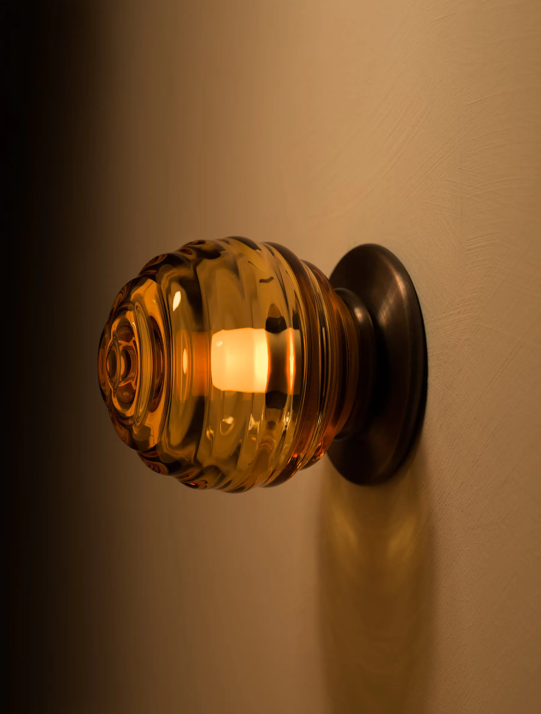 Amber glass spherical wall sconce with ribbed texture and dark bronze mounting bracket against warm beige wall by Allied Maker.