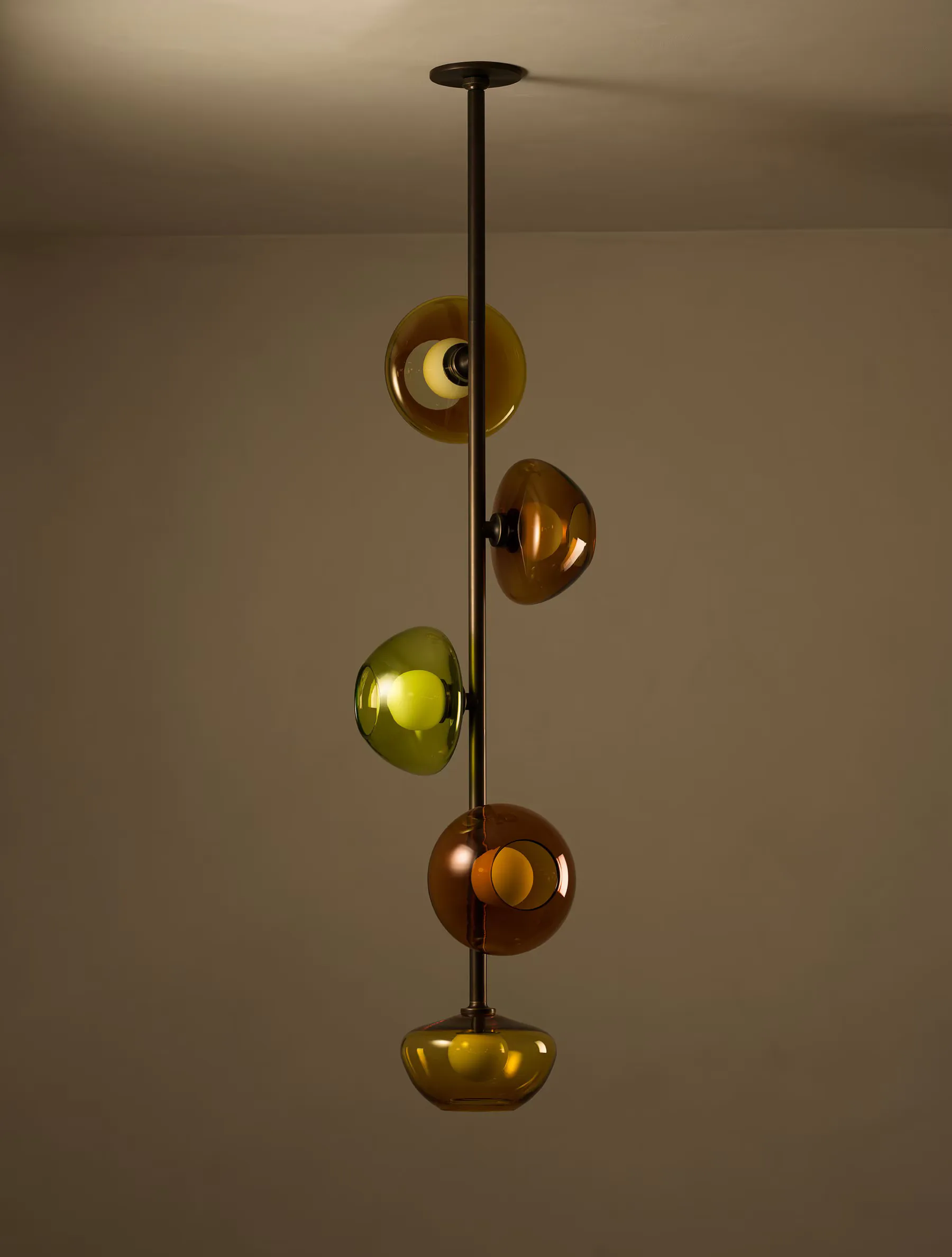 Pendant lighting fixture with five stacked spherical glass globes in warm amber and yellow tones suspended from a bronze rod by Allied Maker.