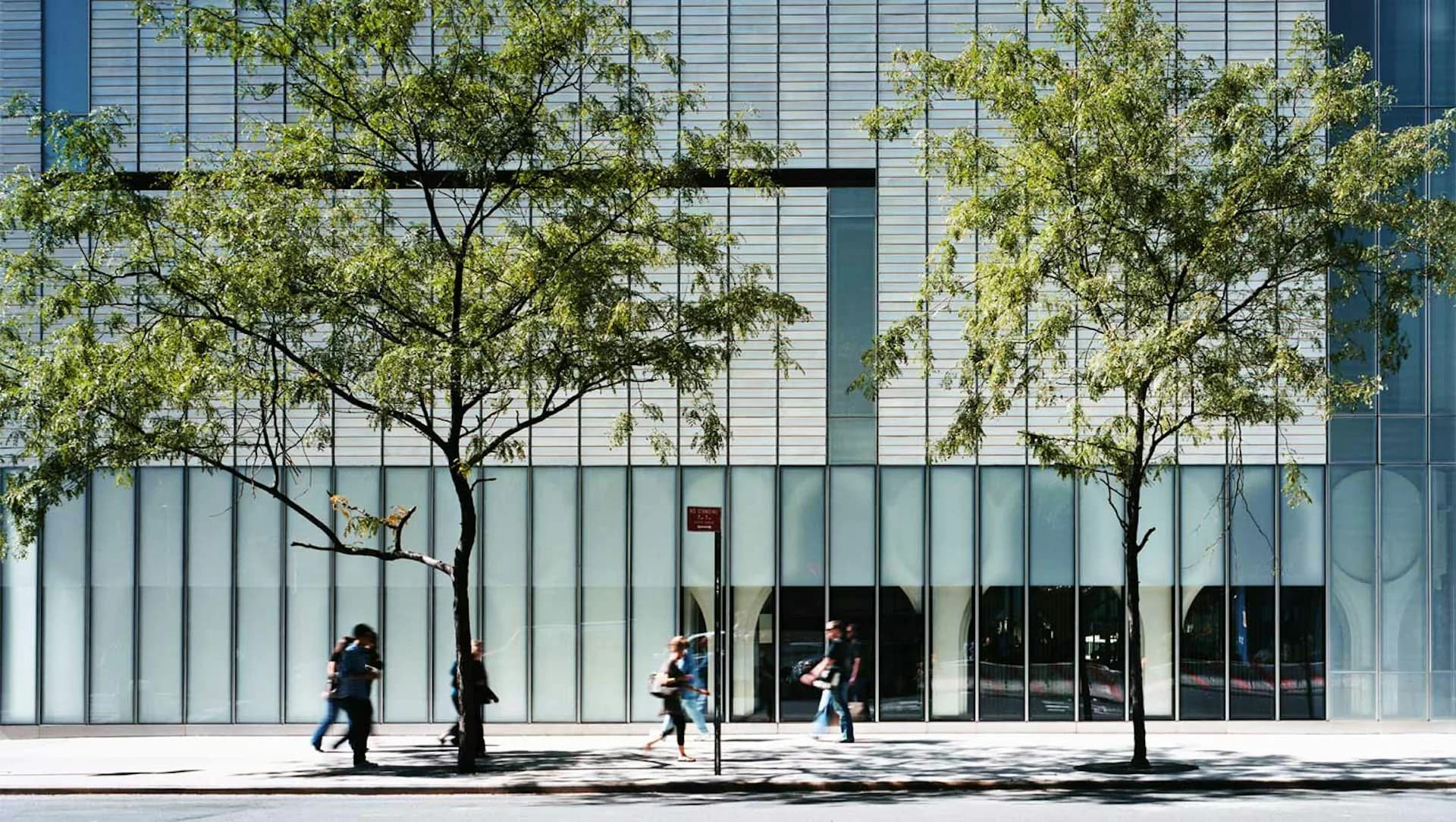 Modern glass-fronted building facade with vertical striations, young trees, and pedestrians walking past, designed by Allied Works.