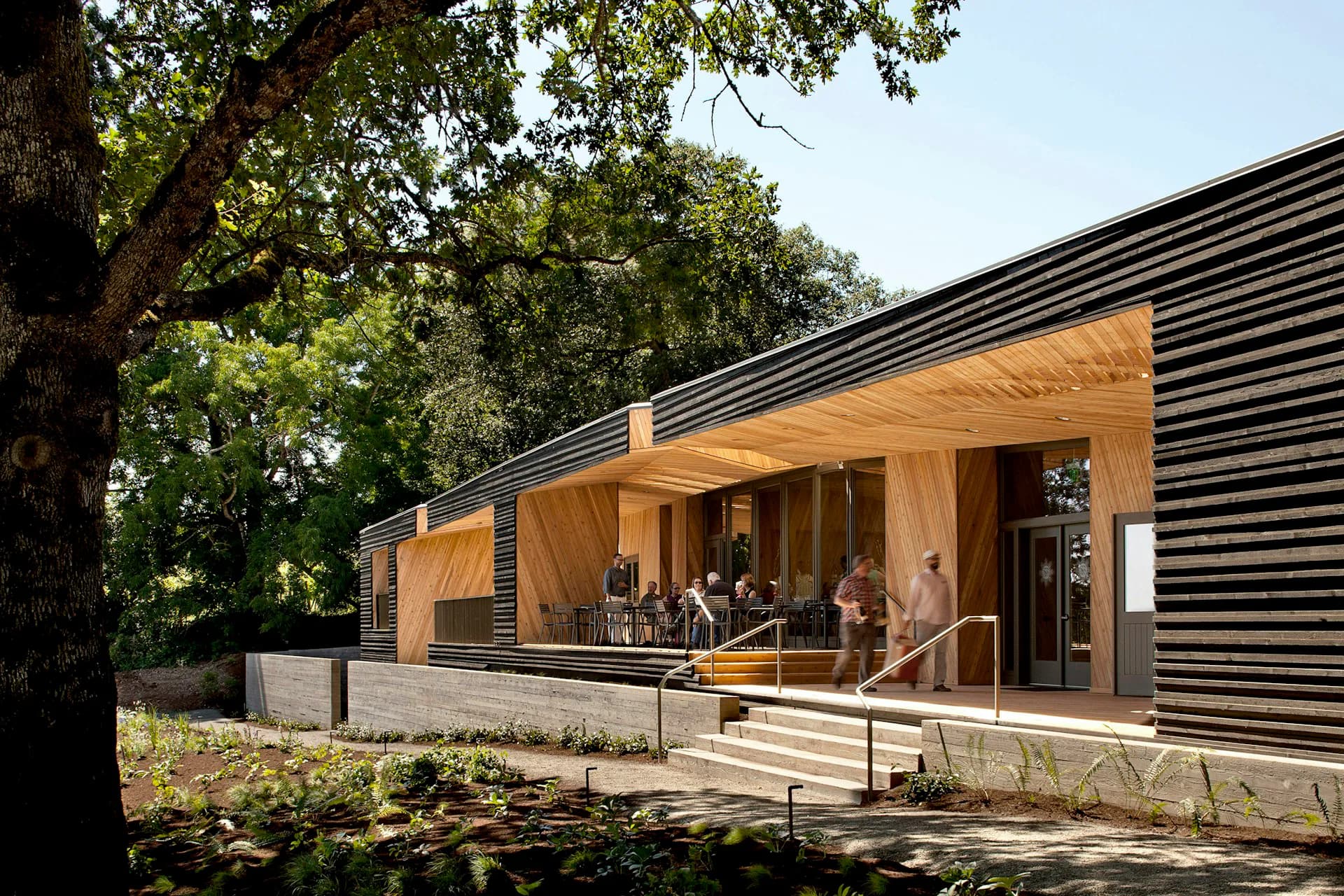 Modern wooden pavilion with black corrugated metal siding, large glazed opening, and concrete base nestled among mature trees by Allied Works.