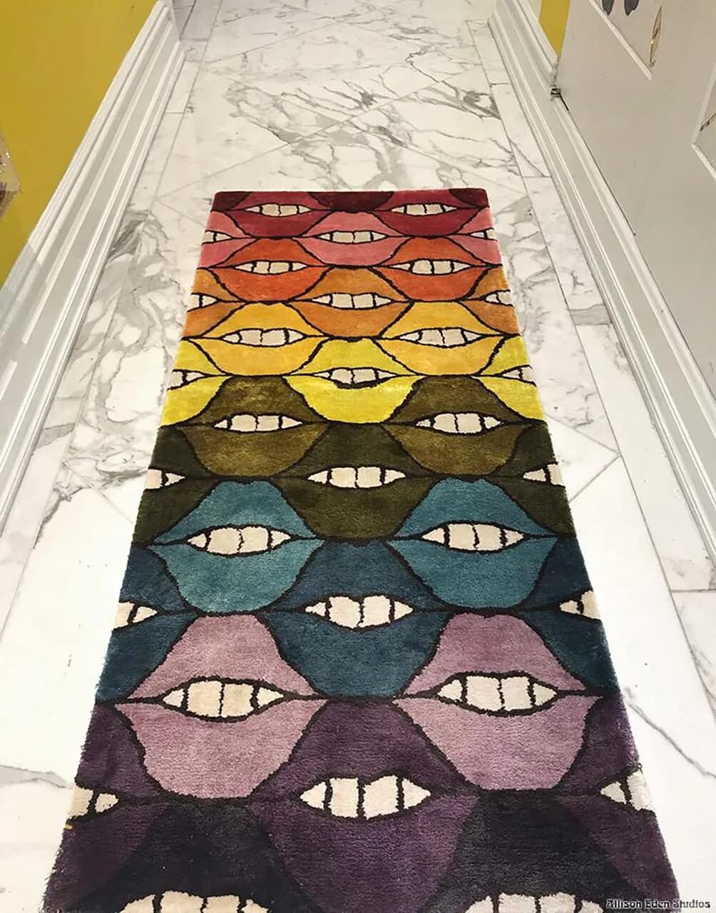 Colorful striped area rug with repeating mouth motif in gradient from burgundy to purple, displayed on marble flooring by Allison Eden Studios.
