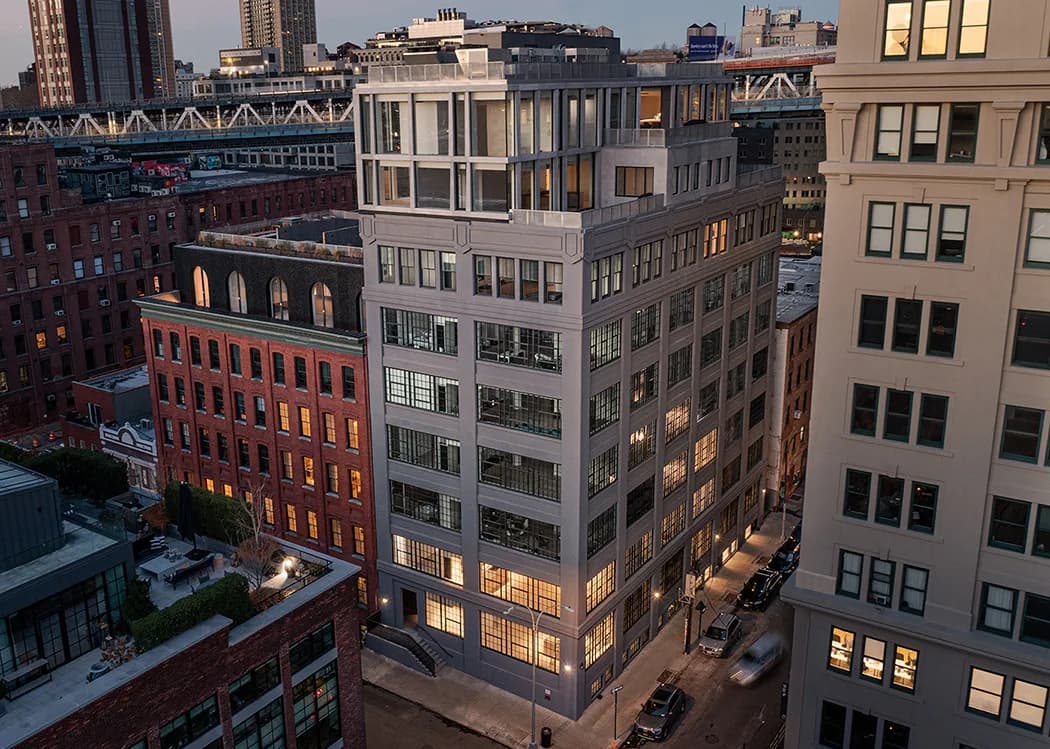 Corner building in urban streetscape at dusk with warm-lit windows, brick and stone facades, adjacent historic structures by Alloy Development.