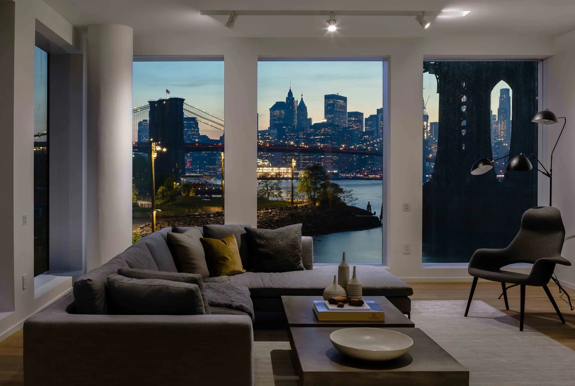Modern living room with charcoal sectional sofa, floor-to-ceiling windows framing Manhattan skyline views, minimalist décor, and contemporary furnishings by Alloy Development.