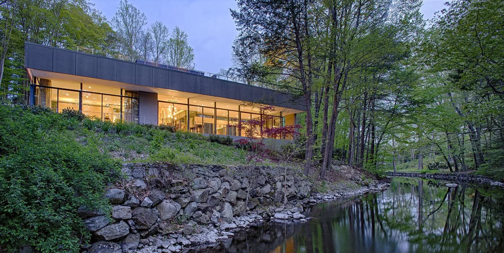 Modern residential building with dark exterior and extensive glazing nestled among trees overlooking a rocky stream, photographed at dusk by Ampersand Architecture.