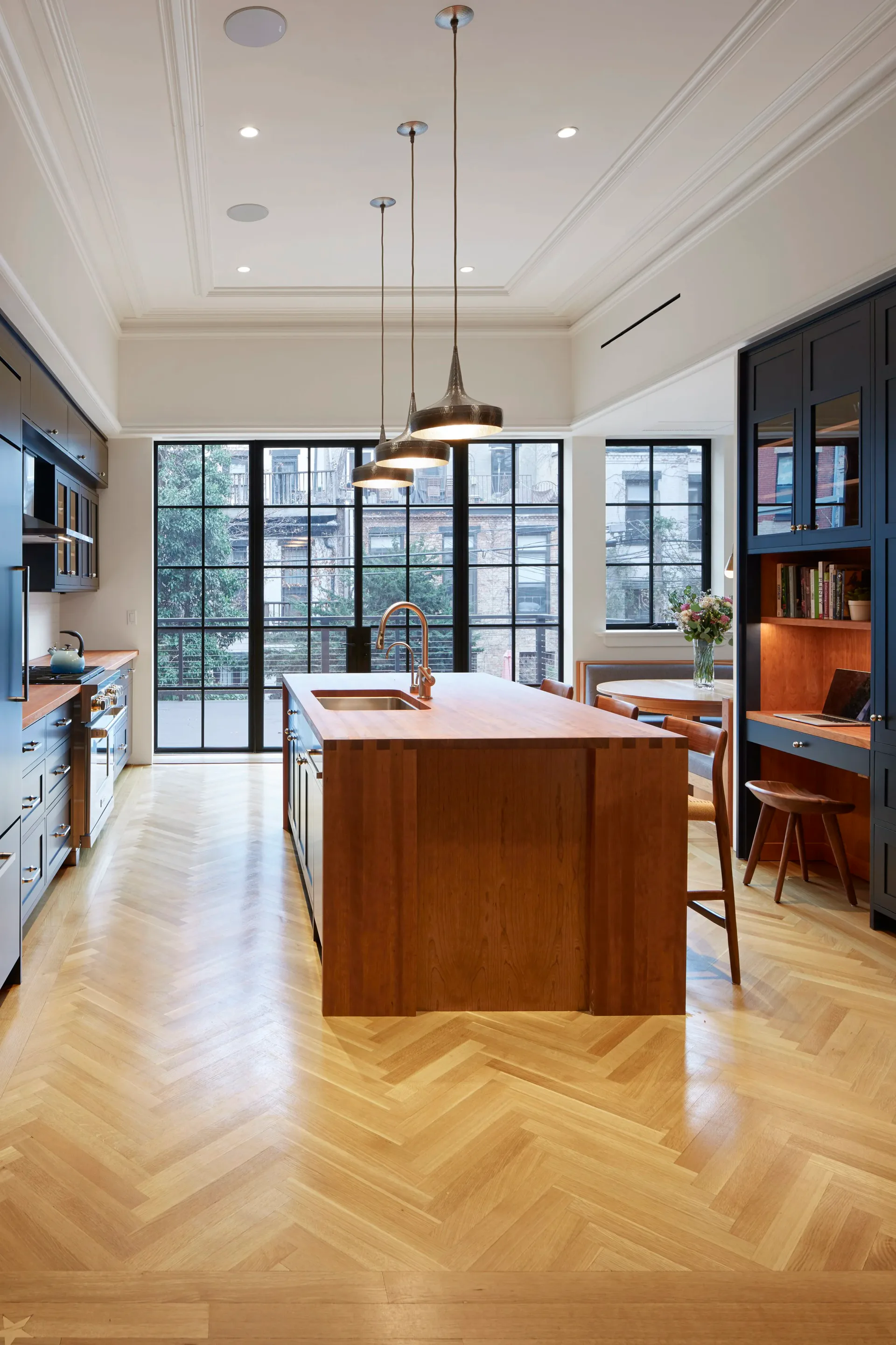 Modern kitchen with walnut island, white cabinetry, herringbone wood flooring, large black-framed windows, and brass pendant lights by Anat Soudry Architect.