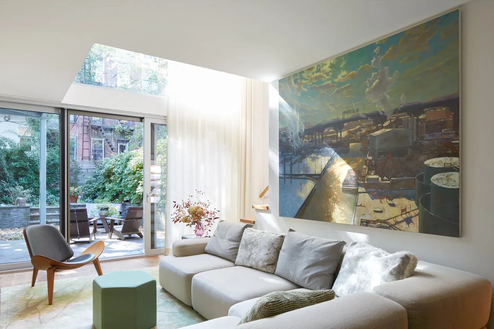 Bright modern living room with light gray sectional sofa, large landscape artwork, floor-to-ceiling windows overlooking a garden, and a sculptural wooden chair by Anat Soudry Architect.