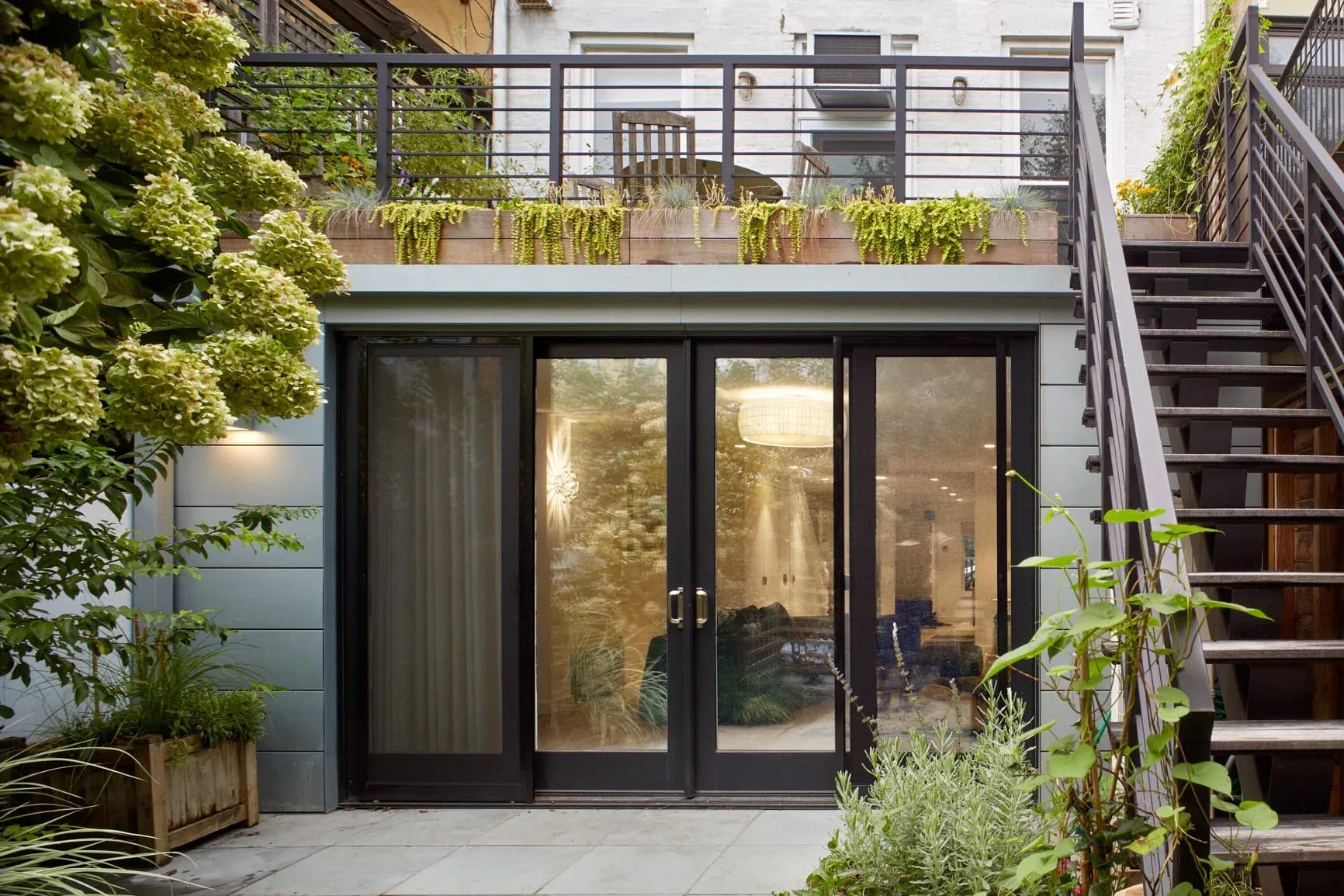 Modern backyard garden room with black-framed glass doors, concrete patio, lush greenery, and steel staircase, designed by Anat Soudry Architect.