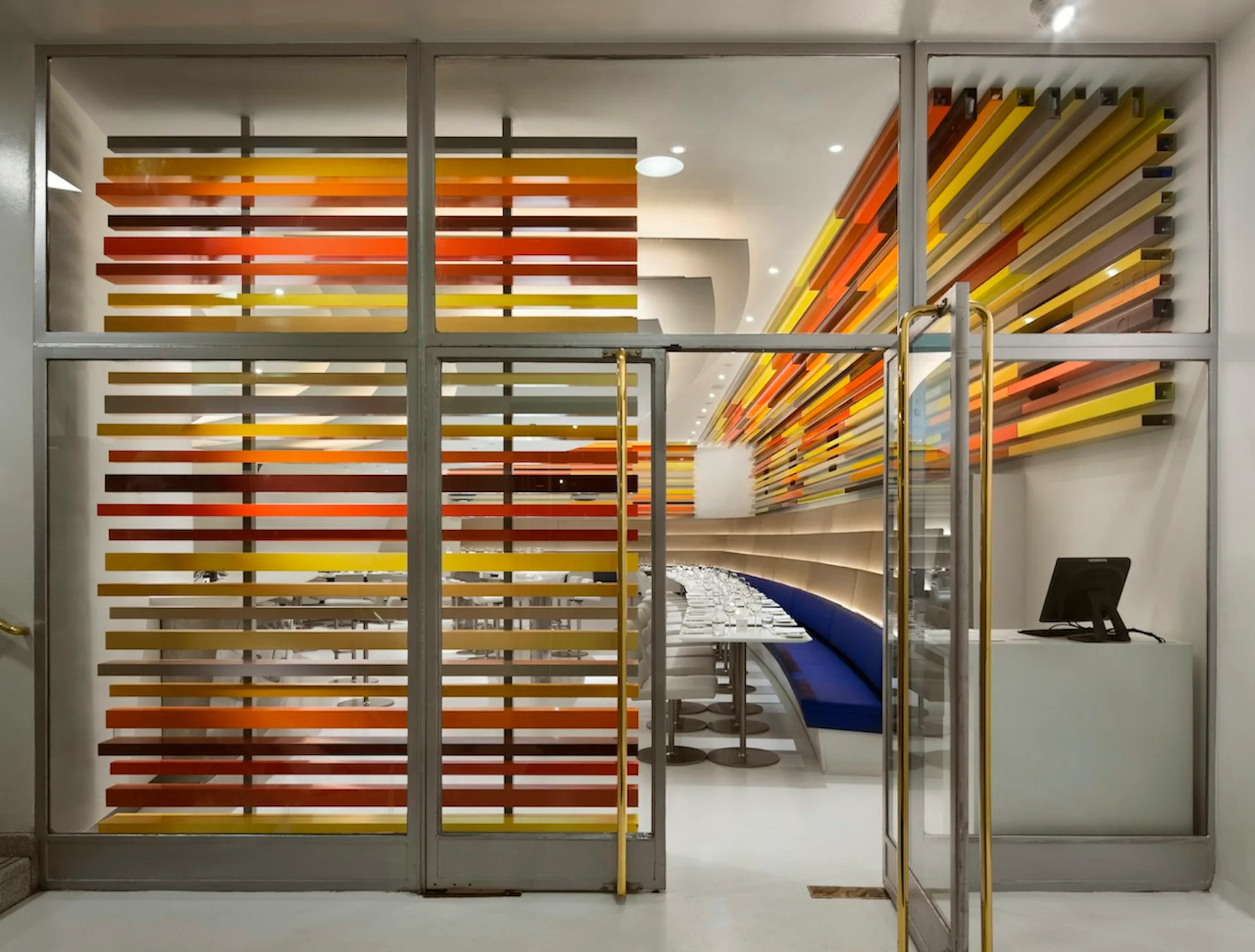 Modern office interior with vibrant horizontal striped wall installation in orange, yellow, and red tones, glass partitions, and minimalist furnishings by Andre Kikoski Architect.