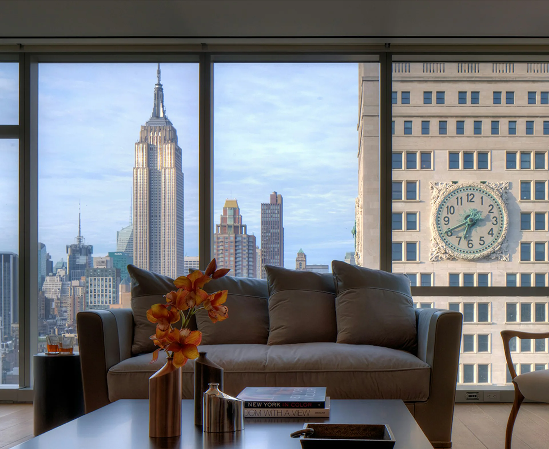 Modern high-rise apartment with floor-to-ceiling windows framing Empire State Building views, charcoal sofa, sunflower arrangement, and minimalist furnishings by Andre Kikoski Architect.