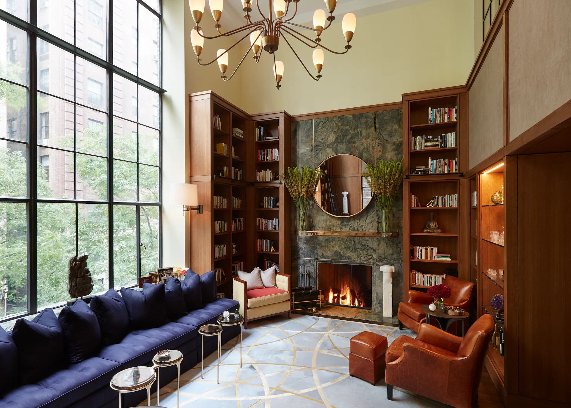 A sophisticated living room with navy blue sectional sofa, burnt orange accent chairs, built-in bookshelves, fireplace with round mirror, and large gridded windows by Andrew B. Ballard, AIA Architecture & Interior Design.