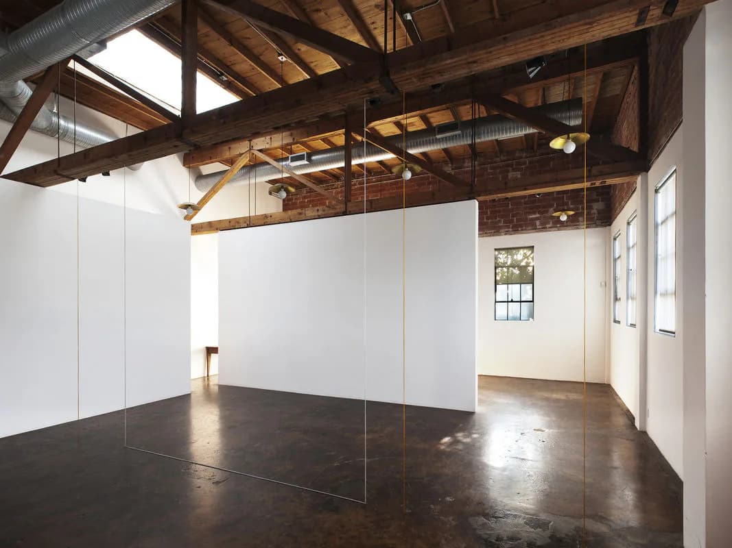 Industrial loft gallery space with exposed wood beams, polished concrete floors, and white glass-walled room, designed by Andrew Berman Architect.