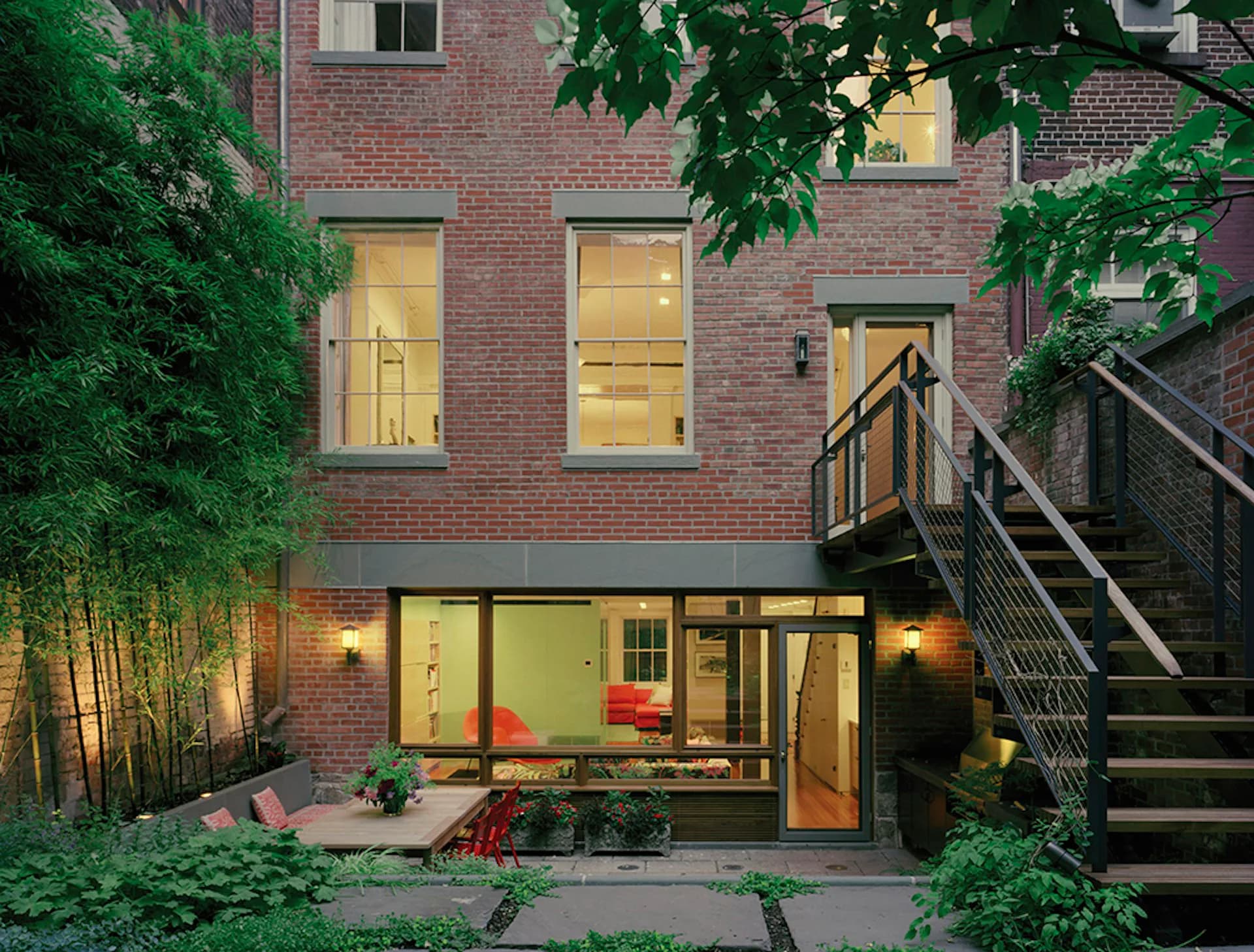 Brick townhouse facade with illuminated ground-floor glass extension, steel staircase, and lush green landscaping by Andrew Franz Architect PLLC.