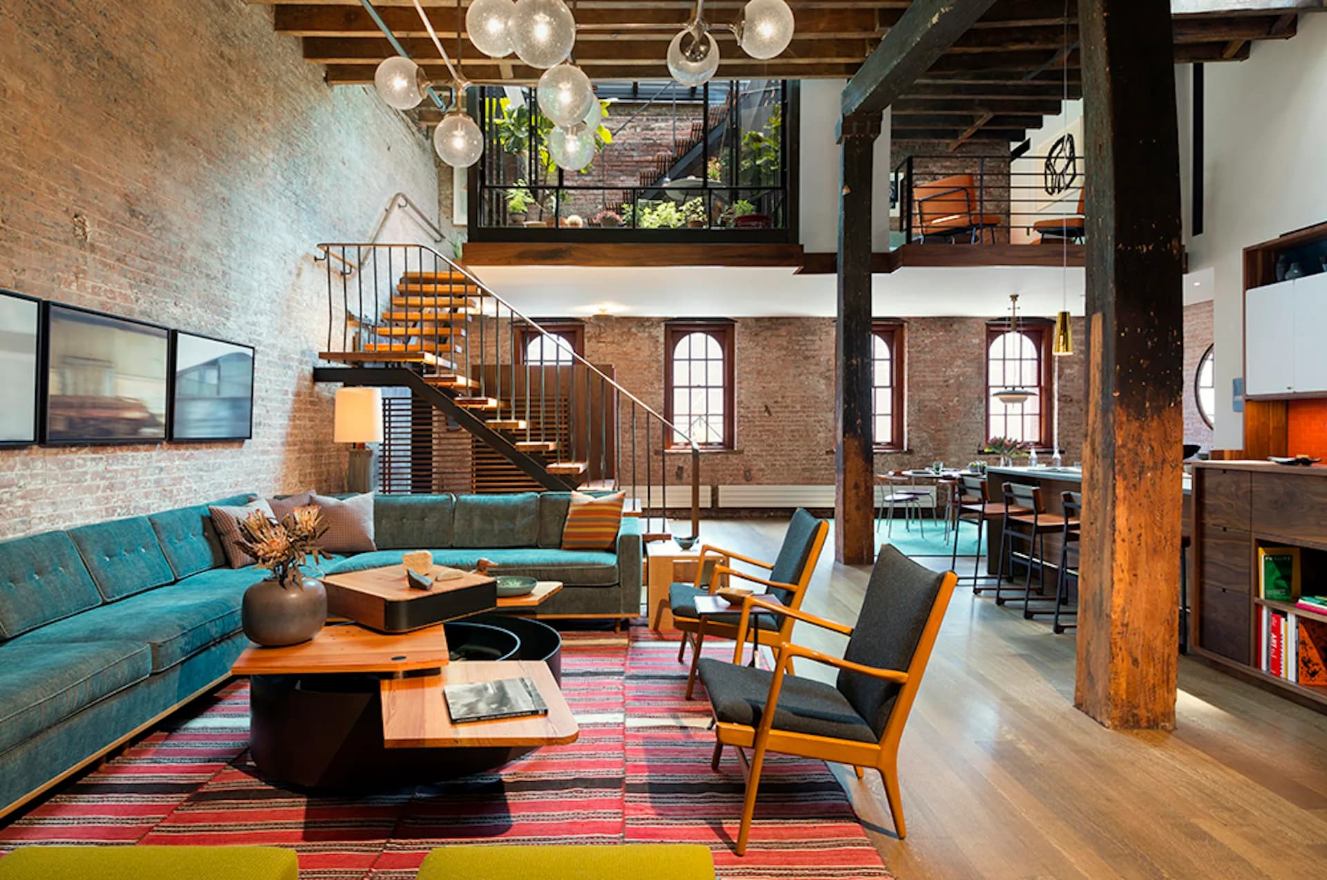 Industrial loft living room with exposed brick, wooden beams, turquoise sofa, mid-century chairs, colorful striped rug, and multi-level mezzanine by Andrew Franz Architect PLLC.
