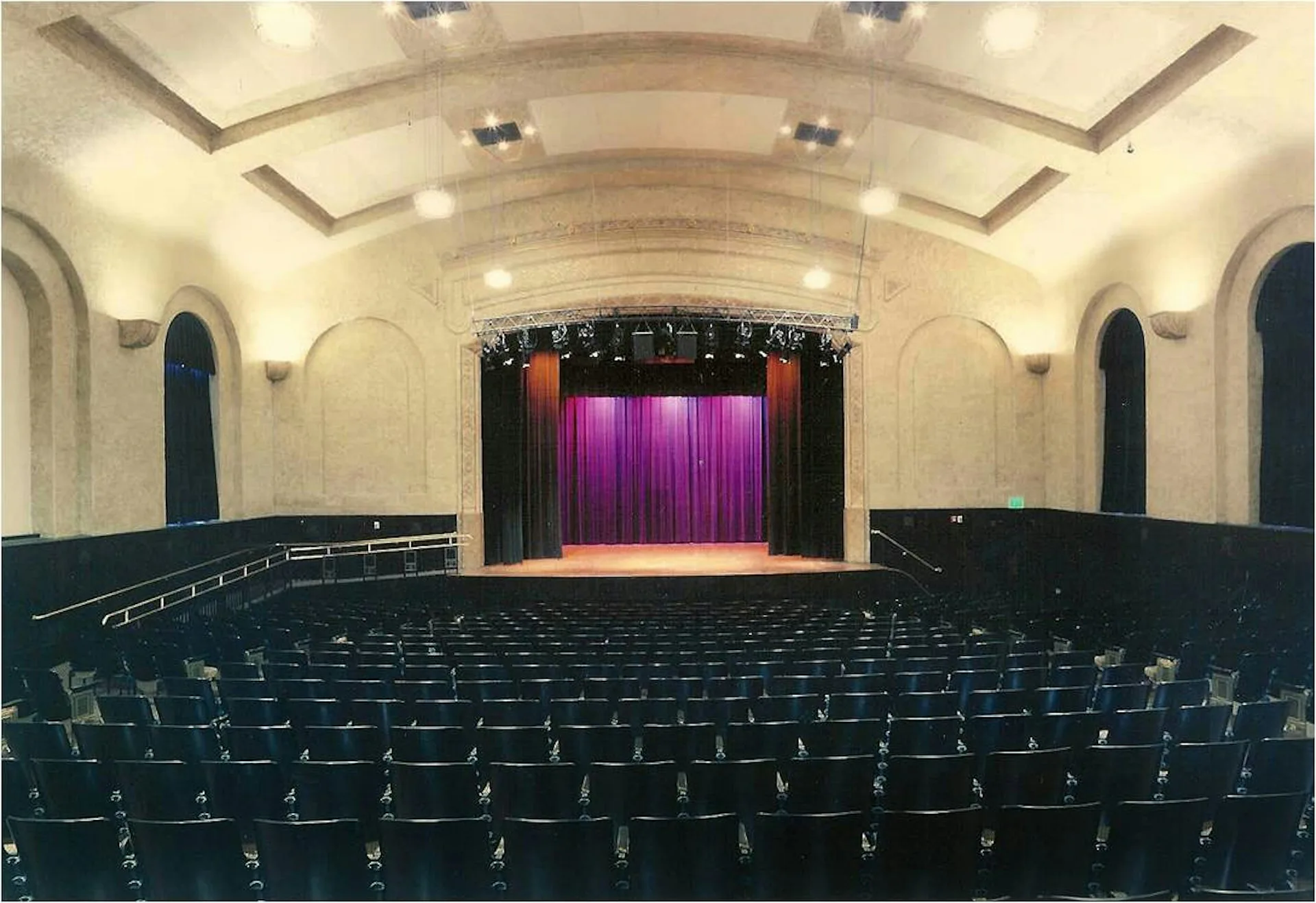 Art deco theater interior with cream coffered ceiling, purple stage curtains, black seating rows, and arched side alcoves by Andrew L. Pettit Architect.