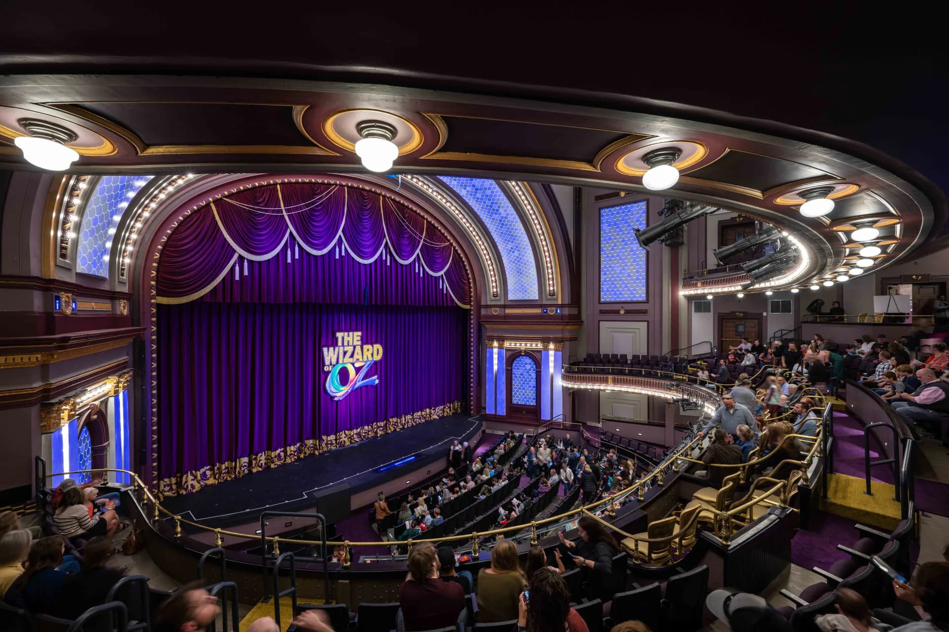 Grand theater interior featuring purple velvet curtains, ornate gold railings, decorative ceiling lights, and seated audience, designed by Apeiro Design.