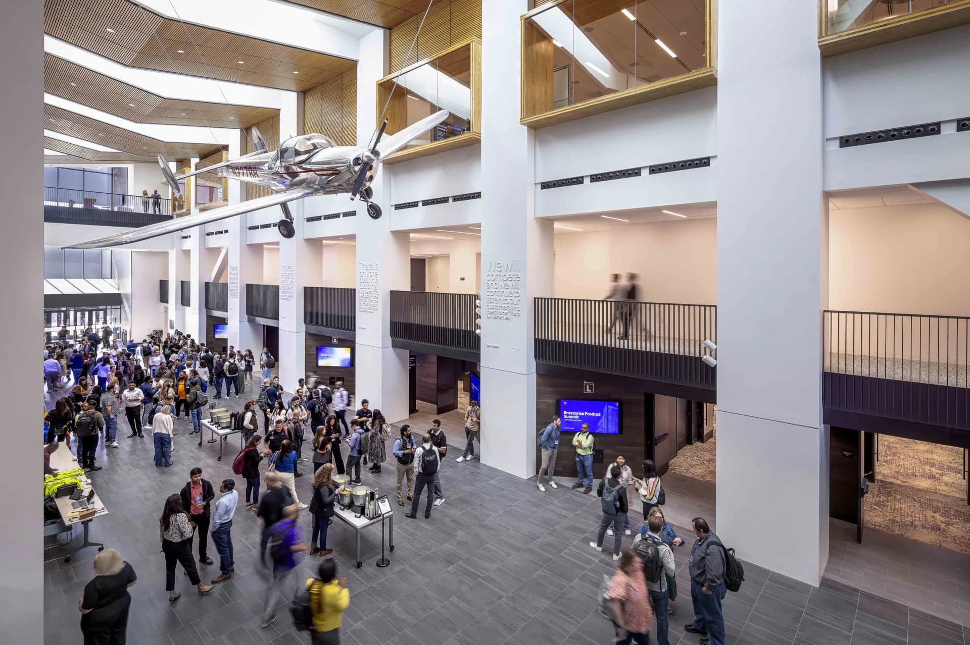 Multi-story public atrium with soaring ceilings, structural columns, skylights, and gathered crowds, showcasing modern interior architecture by Apeiro Design.