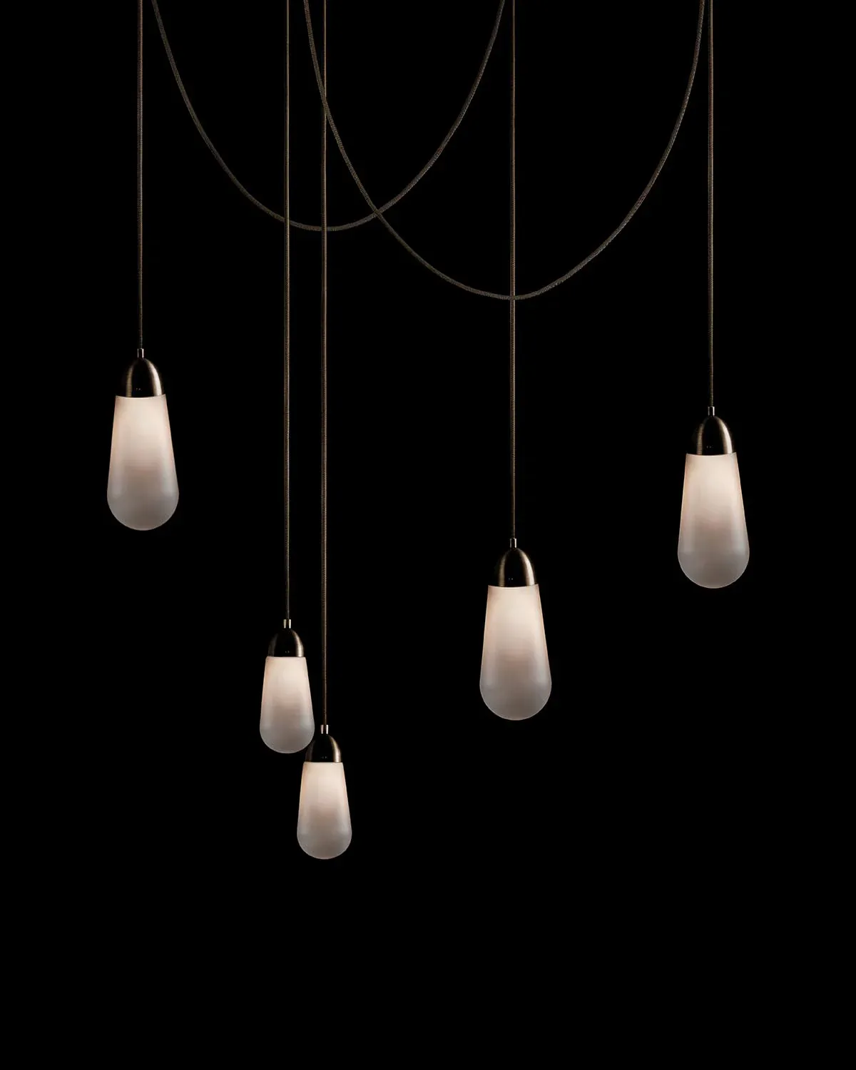 Multiple frosted glass pendant lights suspended from curved cables against a black background, creating an elegant hanging installation by Apparatus.
