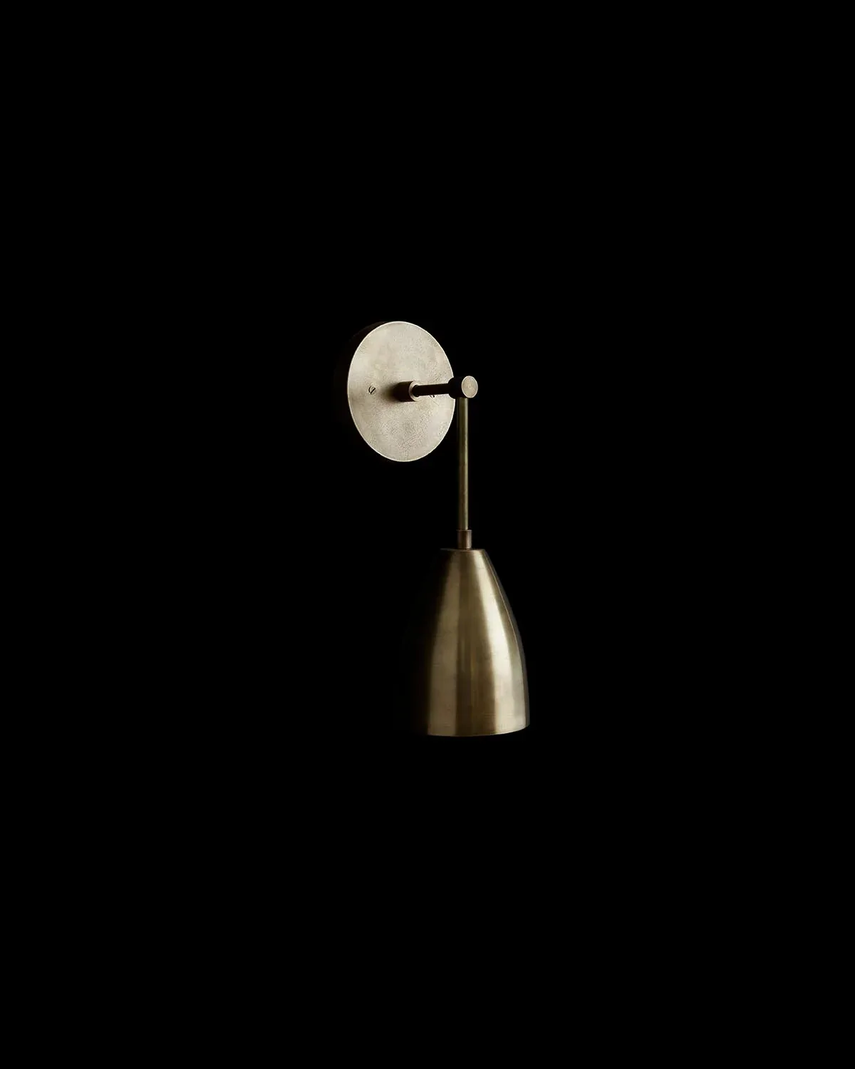 Minimalist brass pendant light with conical shade suspended from circular wall-mounted plate against black background, by Apparatus.