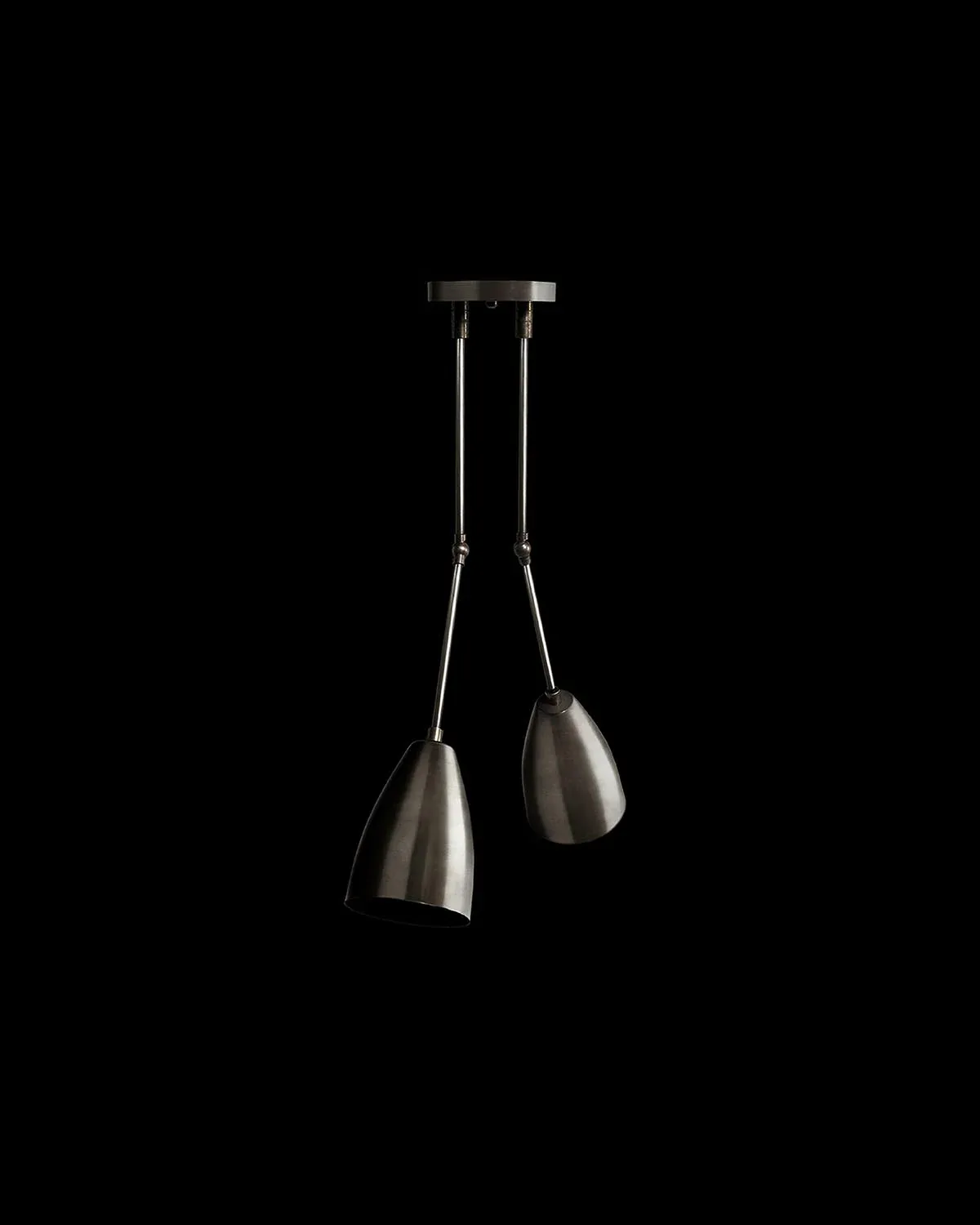 Two pendant lights with brushed metal cone shades suspended from a ceiling-mounted rail system against a black background by Apparatus.