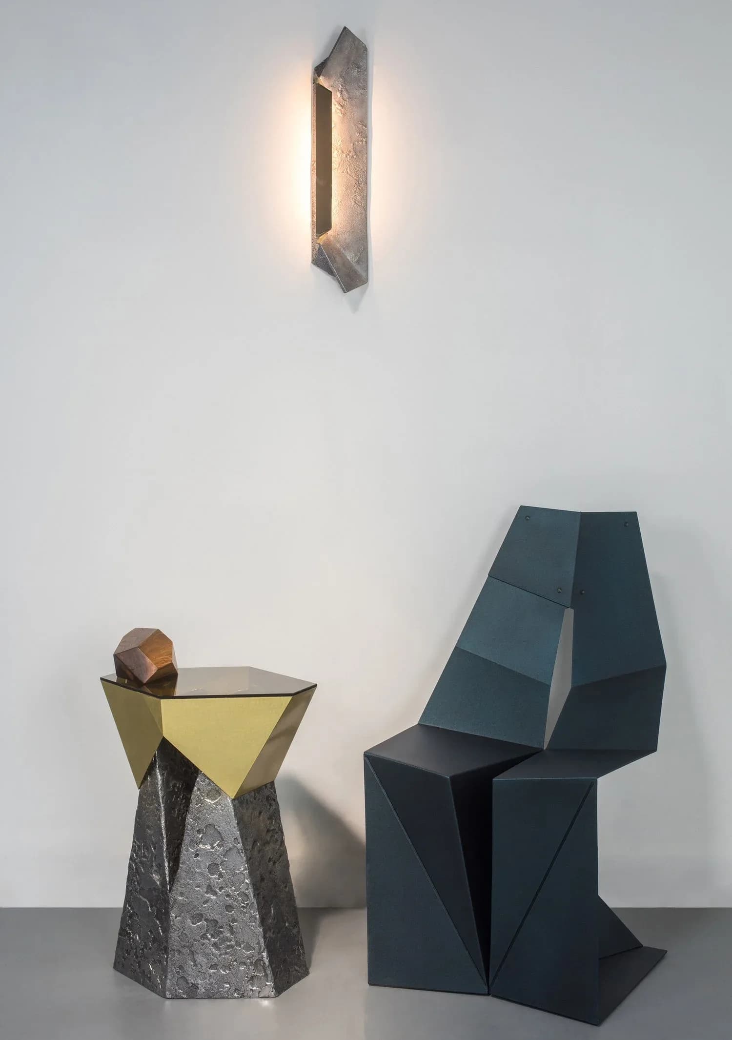 Contemporary installation featuring geometric sculptural forms in dark metal and stone with brass accents, illuminated by a minimalist linear wall sconce by Arcana.