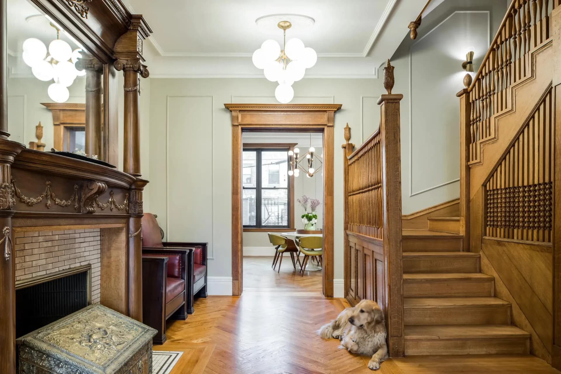 A classical foyer features ornate wooden trim, a fireplace with brick surround, hardwood floors, a grand staircase, and period lighting fixtures by Archetype, LLC.