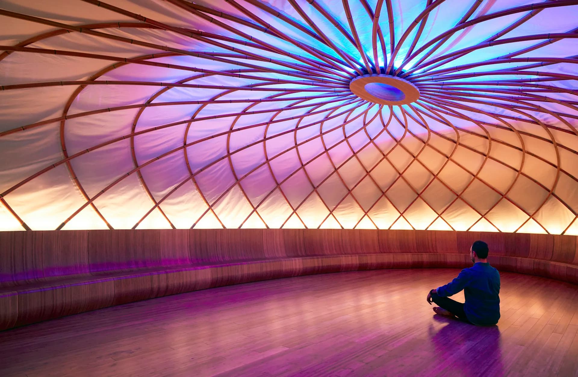 Luminous dome interior with radiating blue and purple lighting, curved ribbed structure, and warm floor surface featuring a seated figure, by Archi-Tectonics NYC, LLC.