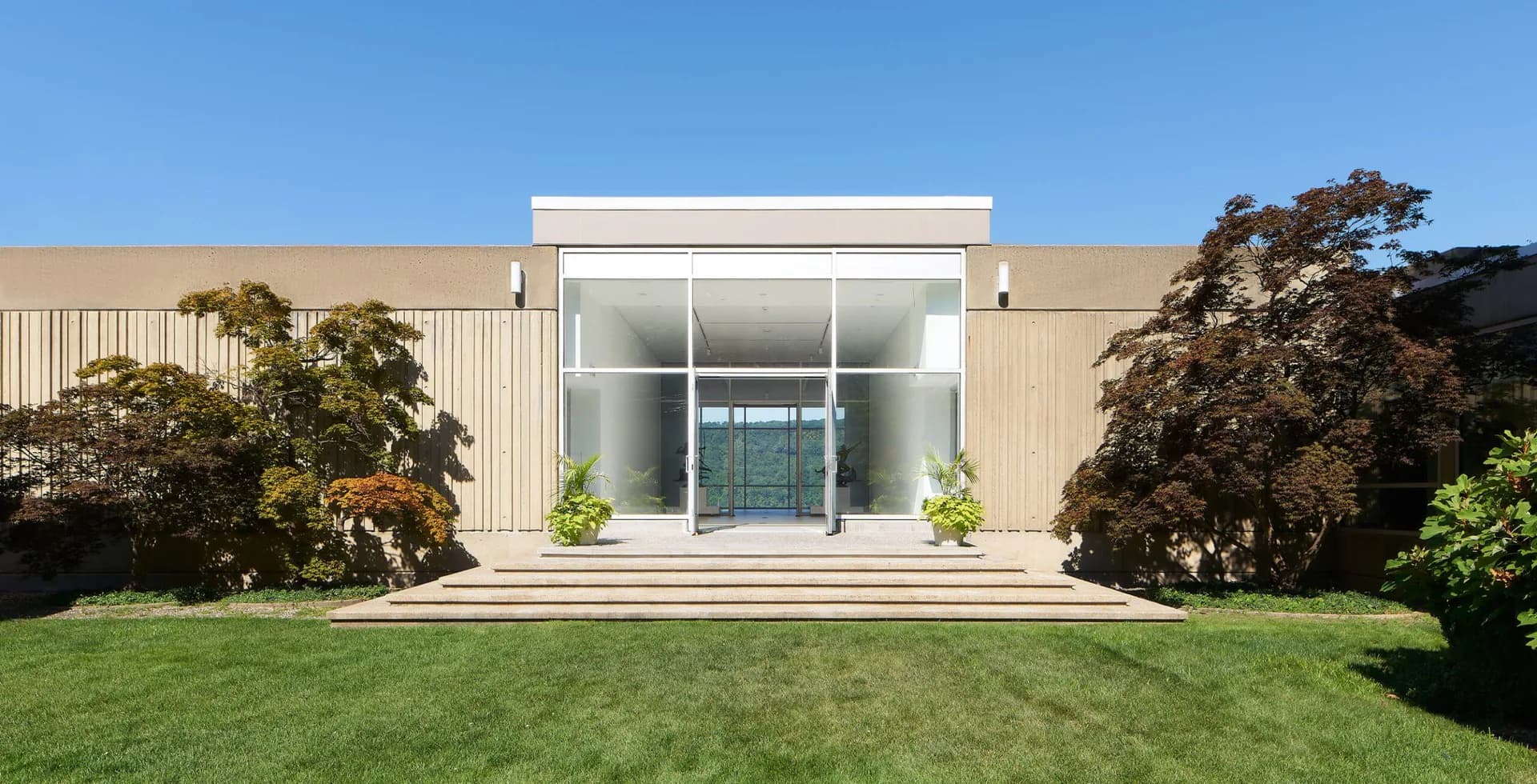 Modern minimalist house with white glass-fronted facade, tan corrugated metal siding, manicured lawn, and symmetrical landscaping by Archimuse.