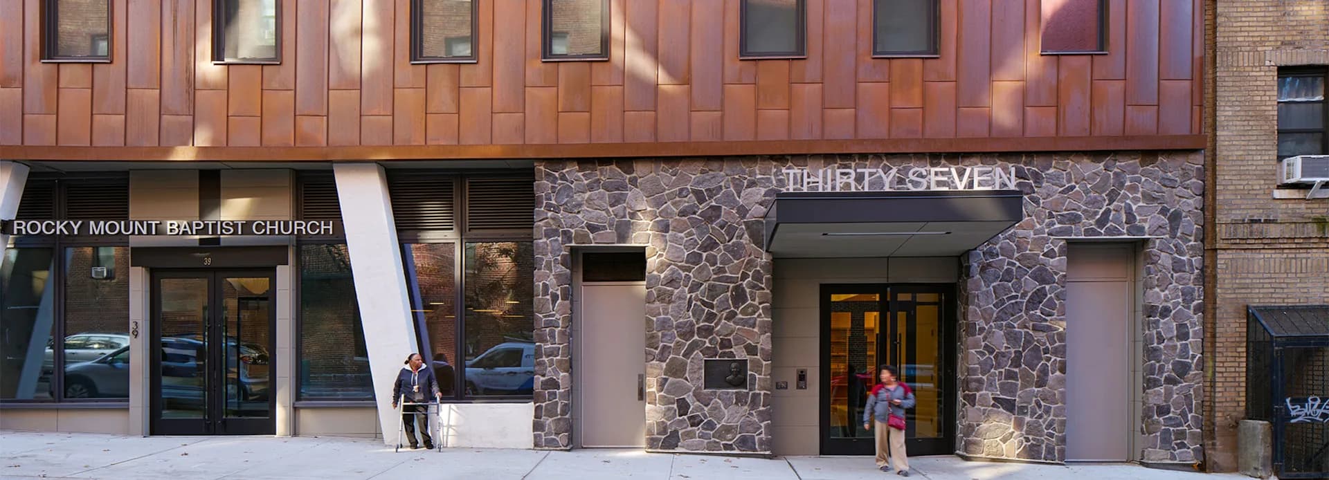 Ground-floor retail storefront featuring warm wood and stone cladding, with modern glass entries and signage for luxury brand tenants by Architecture in Formation.