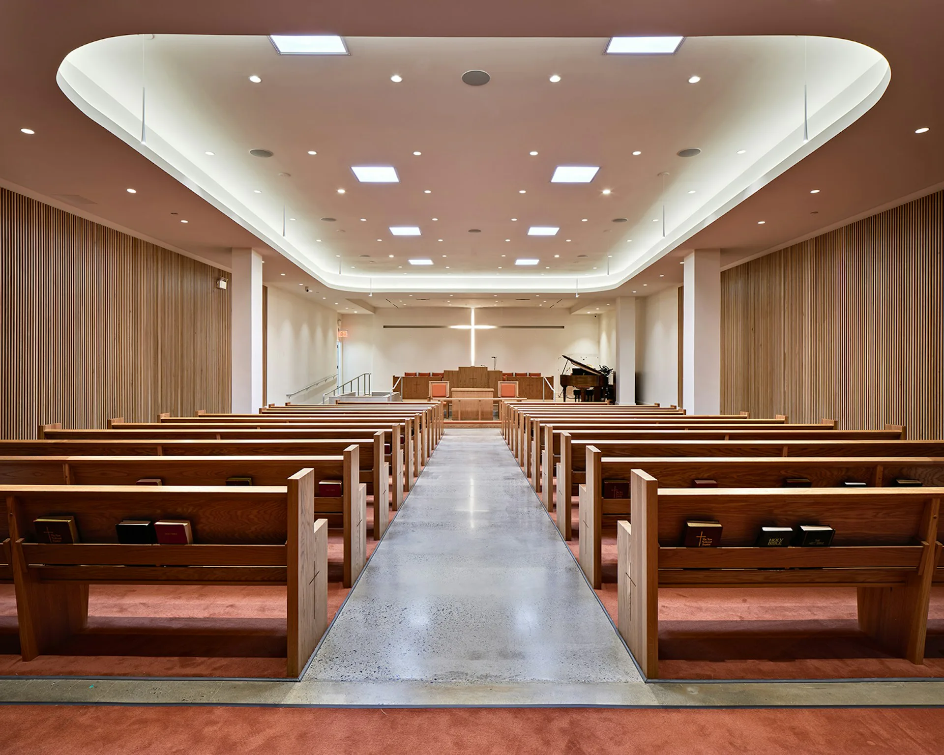 Modern chapel interior with wooden pews, white coffered ceiling with recessed lighting, and altar at far end by Architecture in Formation.