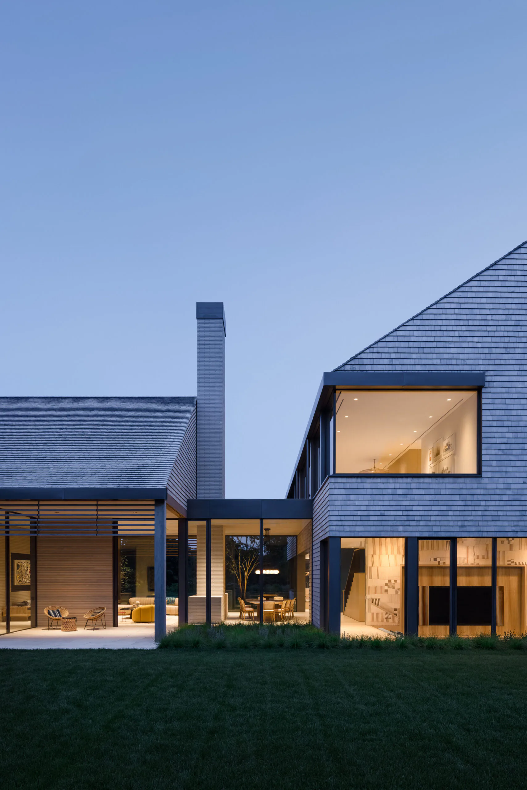 Contemporary residential home at dusk with blue-gray corrugated metal siding, illuminated interior spaces, central chimney, and manicured lawn by ARCHITECTURE OUTFIT PLLC.