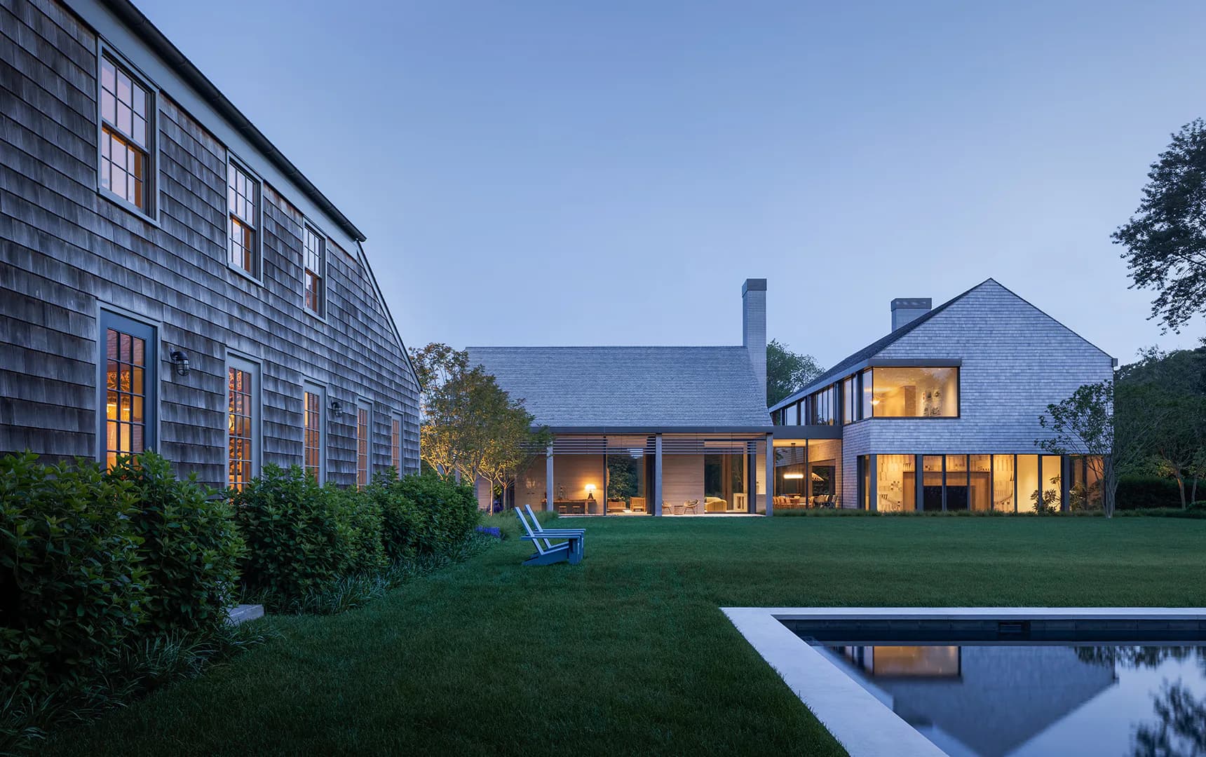 Modern residential exterior at dusk featuring a dark-shingled house with illuminated windows and a clean-lined addition, lush lawn, and rectangular reflecting pool by ARCHITECTURE OUTFIT PLLC.