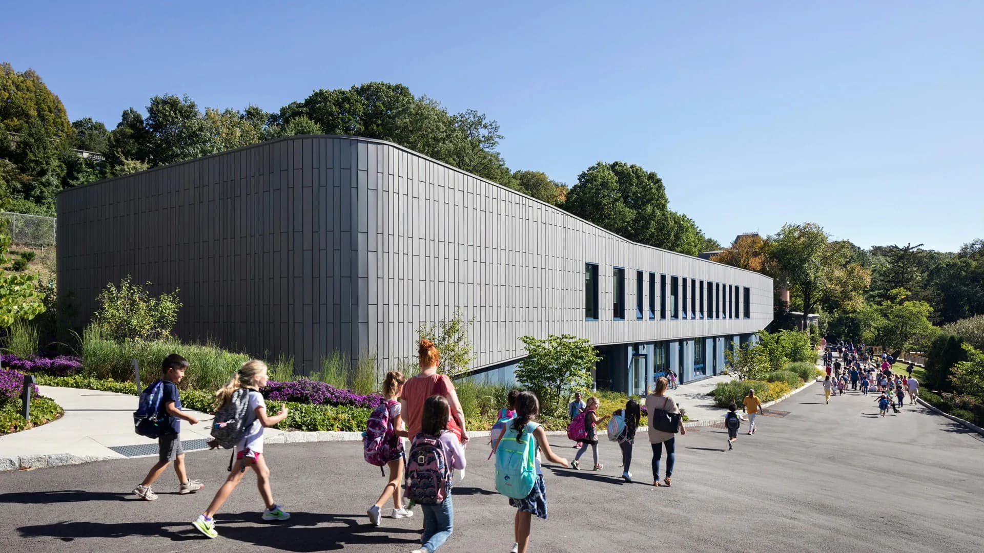 Modern institutional building with dark corrugated metal facade and horizontal window bands, surrounded by trees with people on the plaza forecourt, by Architecture Research Office.