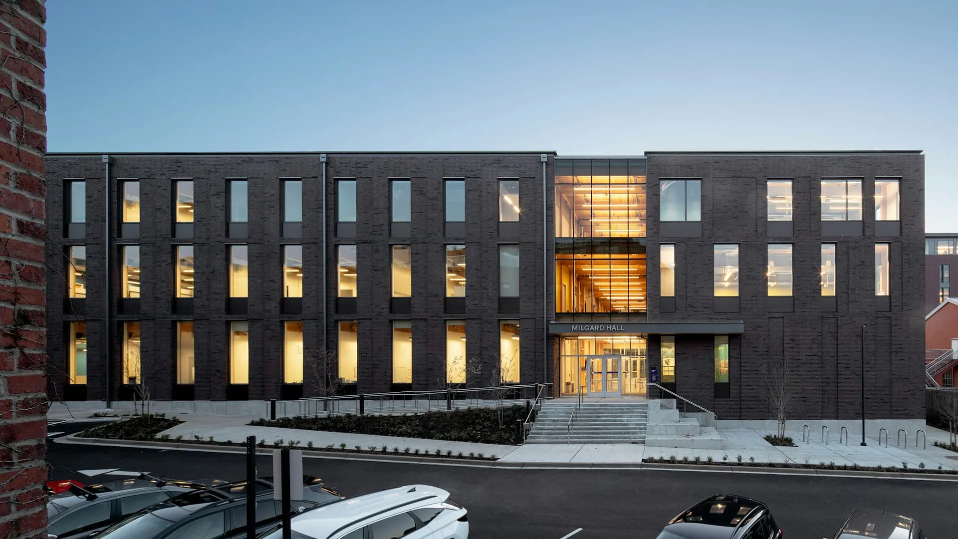 Modern dark-clad building with golden-lit window openings at dusk, featuring geometric facade and minimalist landscaping by Architecture Research Office.