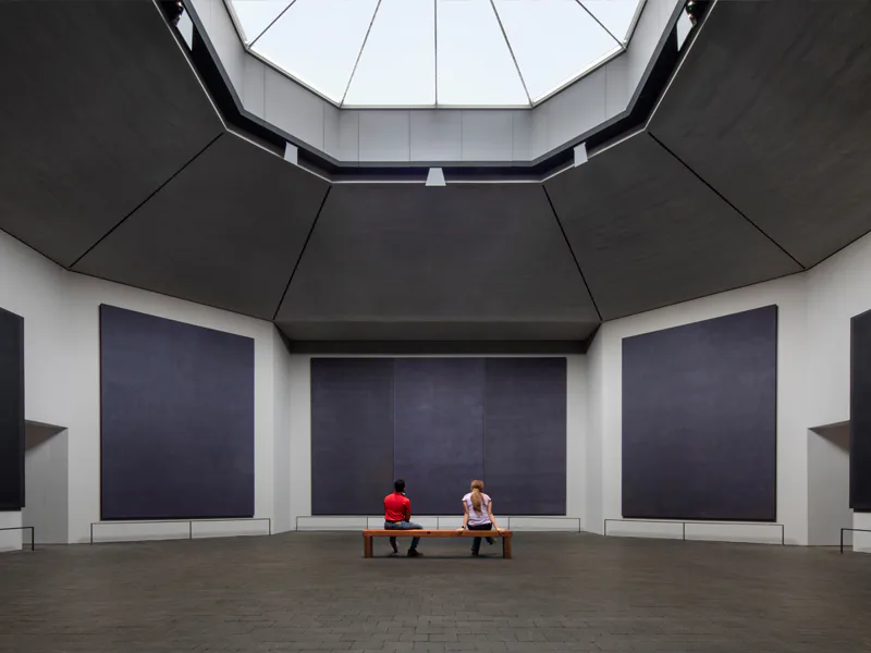A minimalist gallery interior with octagonal skylight, dark ceiling panels, white walls, and dark display panels, featuring two visitors seated on benches by Architecture Research Office (ARO).