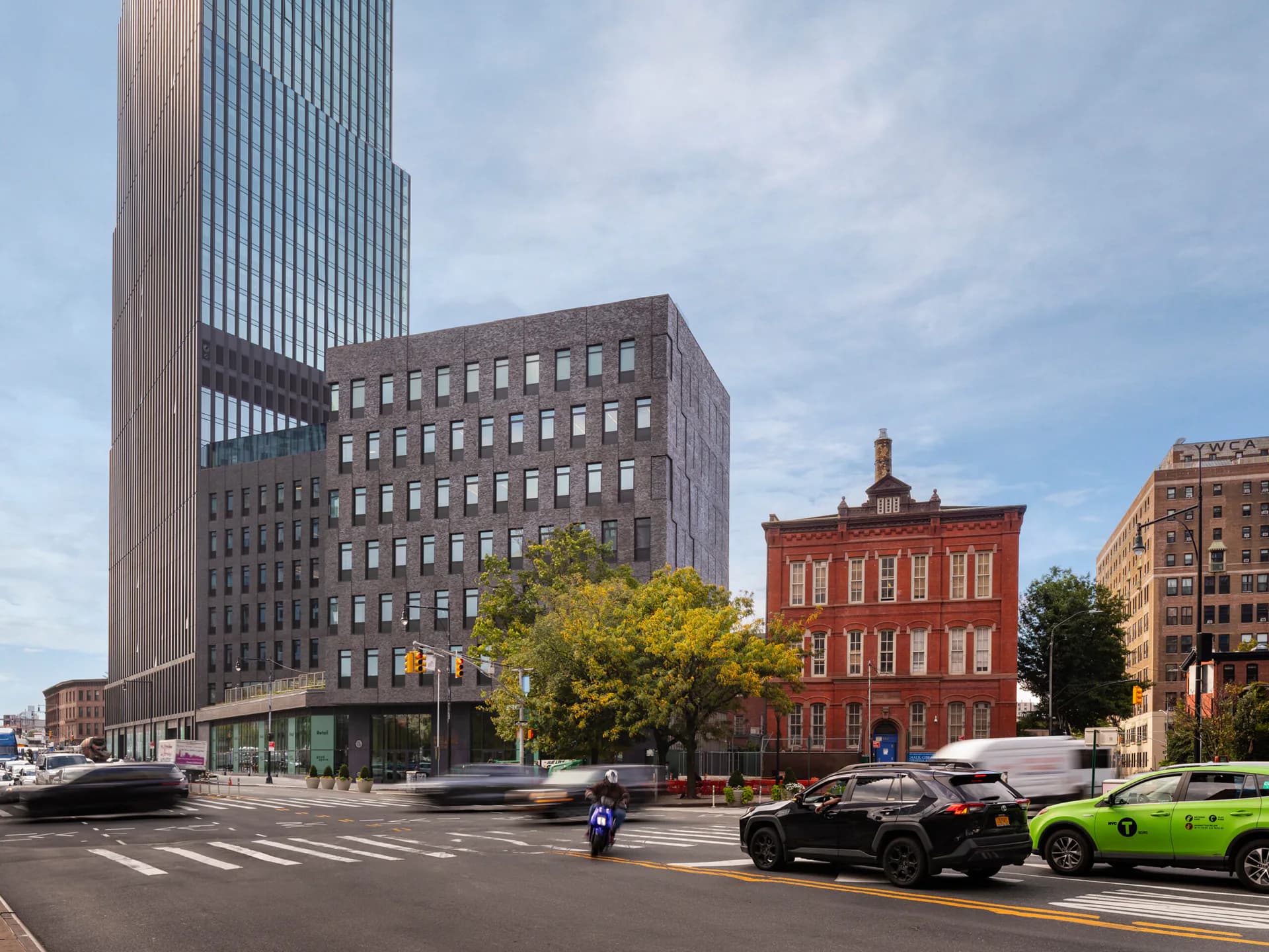 Urban street corner featuring a modernist tower with vertical ribbing alongside a restored red brick historic building, mature trees, and contemporary street-level activity by Architecture Research Office (ARO).