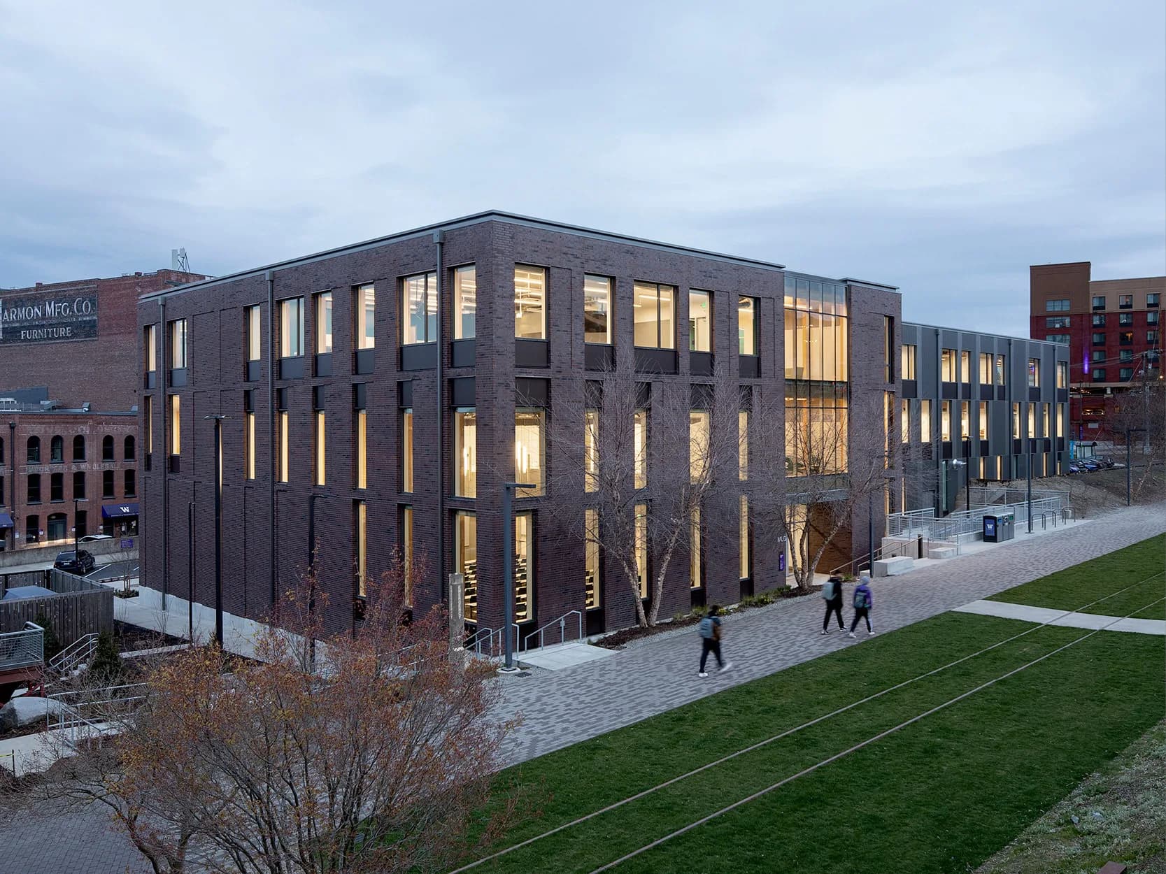 Modern dark brick building with illuminated rectangular windows at dusk, surrounded by green lawn and plaza, by Architecture Research Office (ARO).