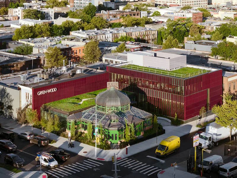 Aerial view of a mixed-use building with distinctive red corrugated metal facades, green rooftop gardens, and a central greenhouse structure surrounded by urban streetscape by Architecture Research Office (ARO).