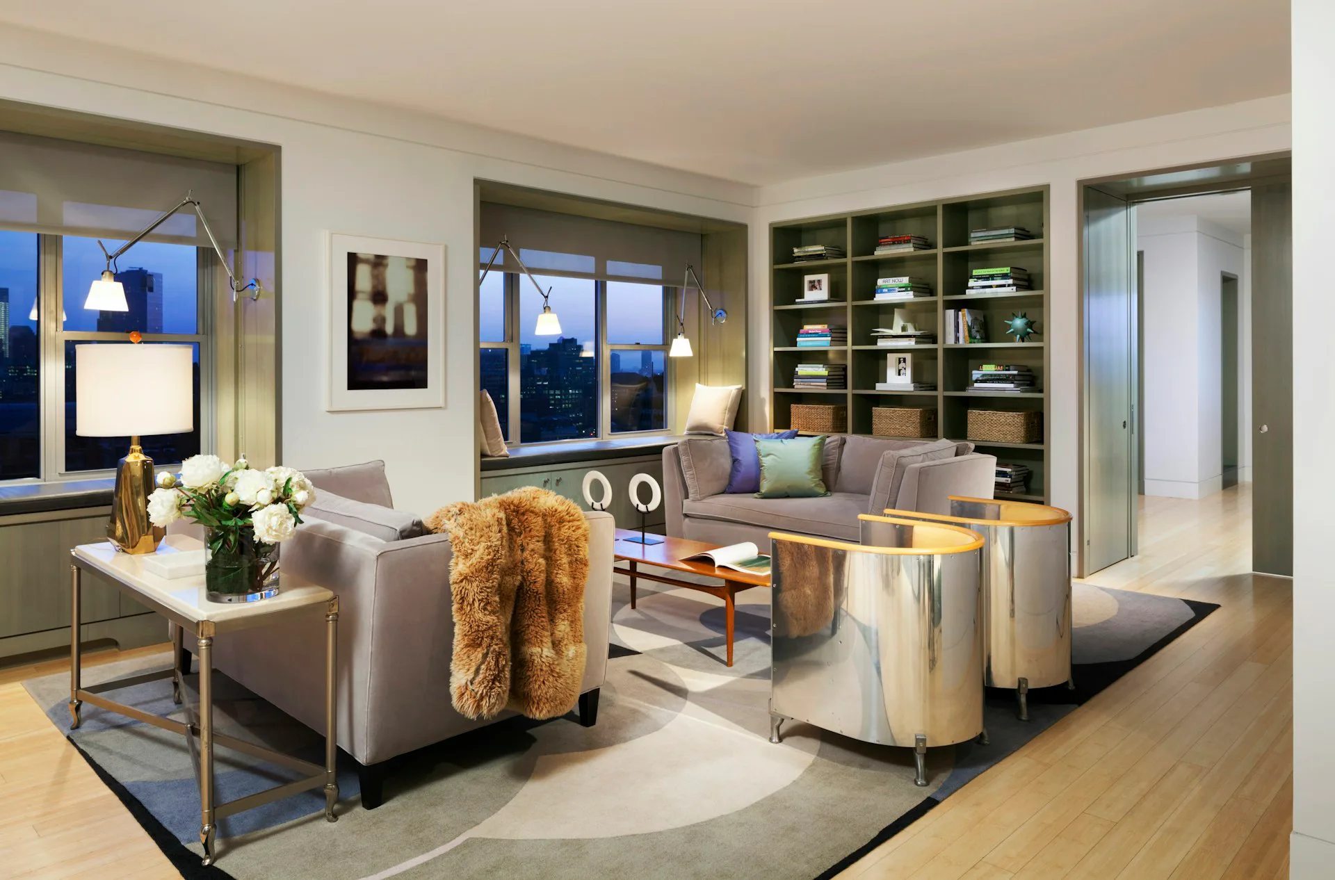 Luxurious modern living room with metallic accent chairs, built-in shelving, floor-to-ceiling windows, and warm wood flooring, designed by Armand LeGardeur Architect.