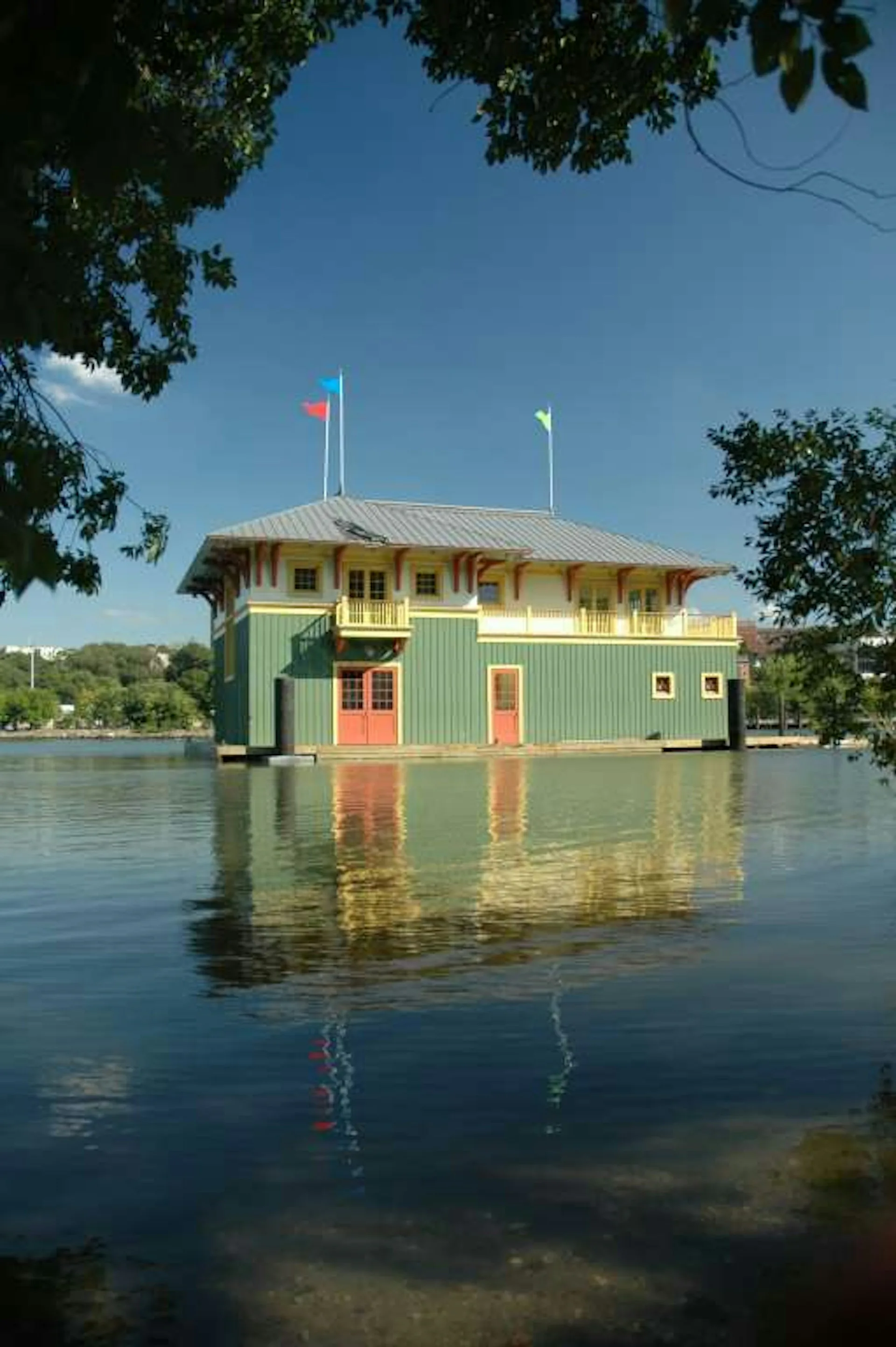 Colorful waterfront boathouse with yellow upper level and green corrugated metal base, reflected in calm water, designed by Armand LeGardeur Architect.