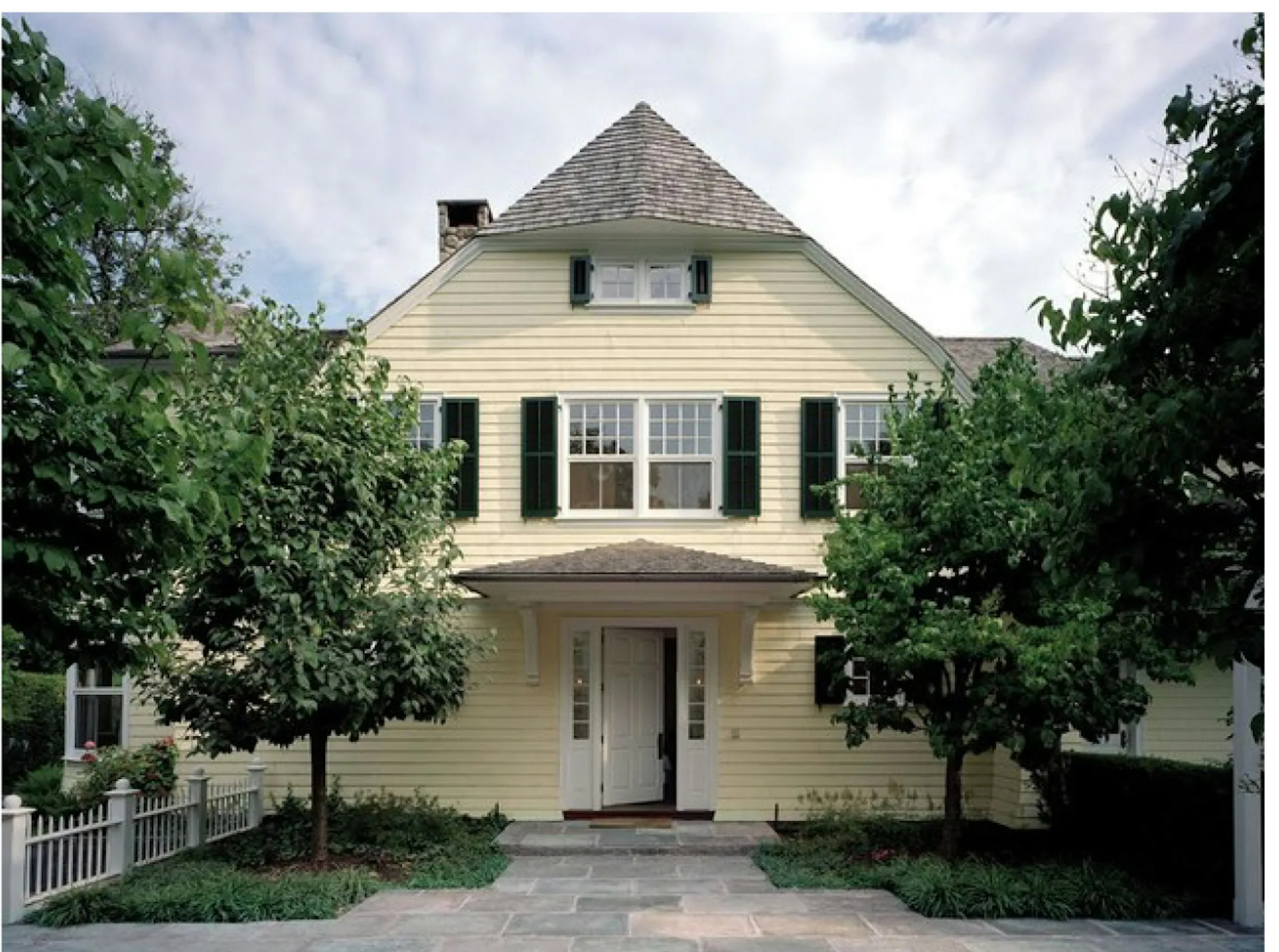 A cream-colored colonial-style residential facade with a peaked roof, black shutters, and symmetrical front entrance framed by mature landscaping, designed by Armand LeGardeur Architect.