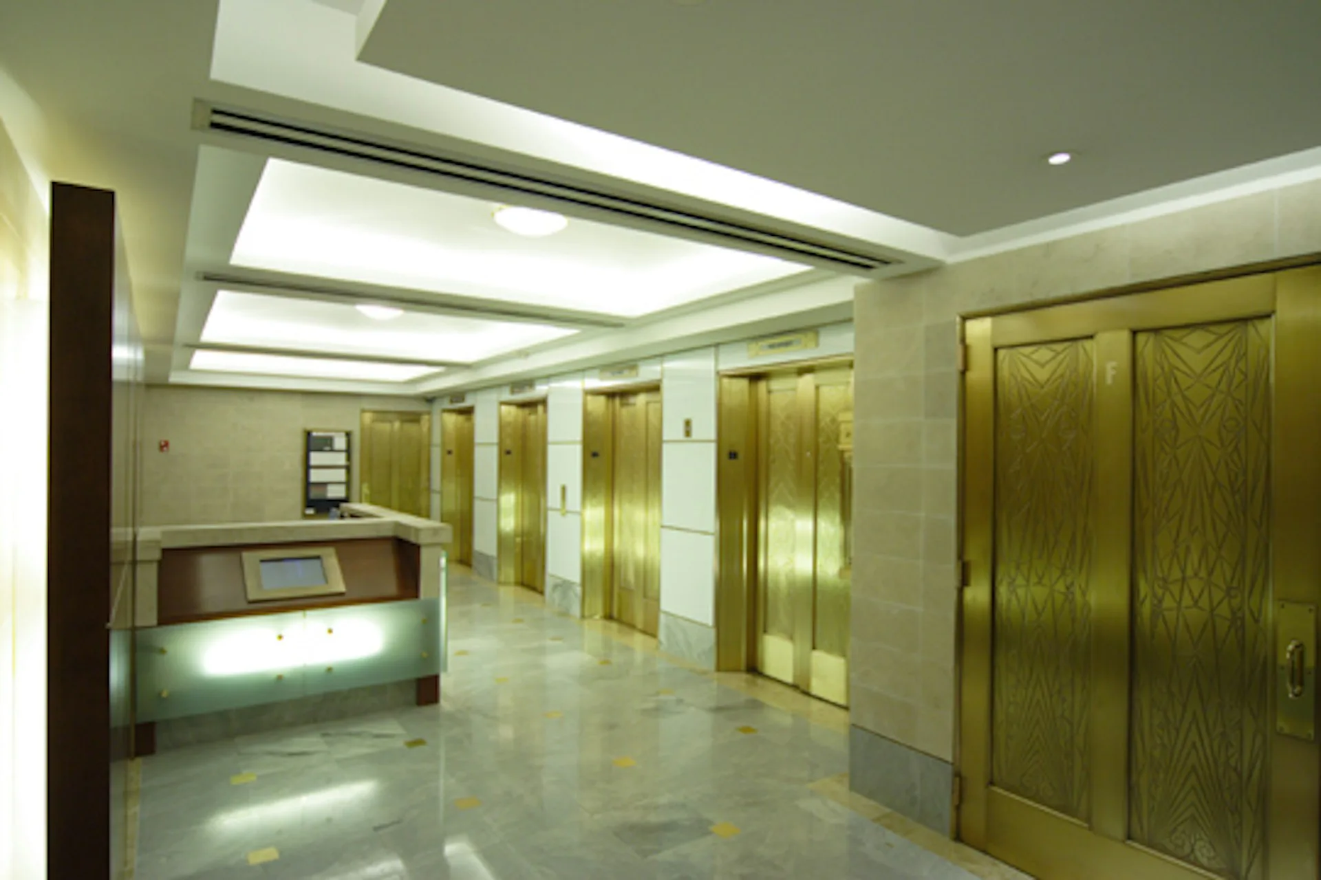Modern elevator lobby with polished marble floors, brass-finished elevator doors, recessed ceiling panels with integrated lighting, and a reception desk, designed by Arpad Baksa Architect, P.C.