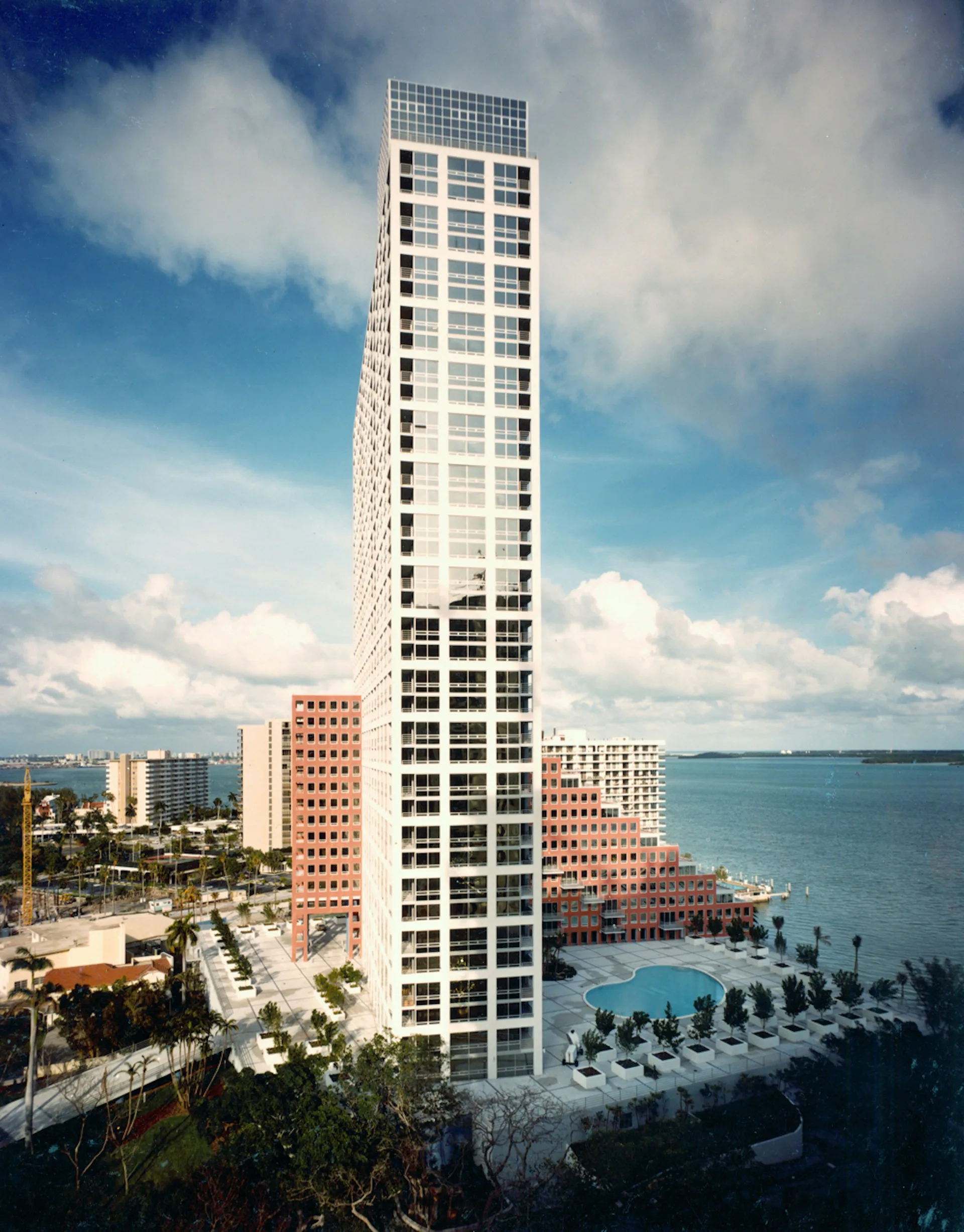 Tall white residential tower with gridded facade overlooking a waterfront plaza with pool and palm trees, by Arquitectonica.