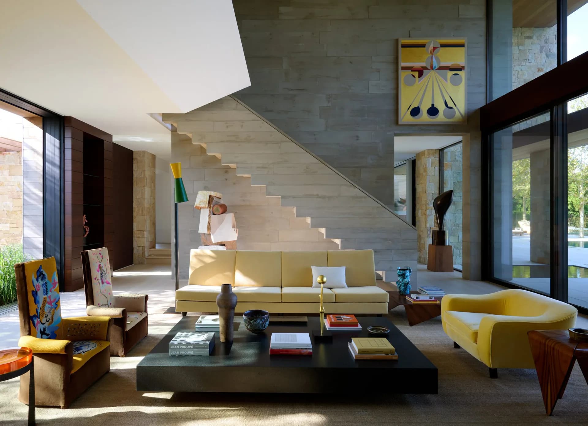Modern living space with concrete curved staircase, yellow upholstered seating, black coffee table, and floor-to-ceiling glazing overlooking landscape, designed by Ashe Leandro.
