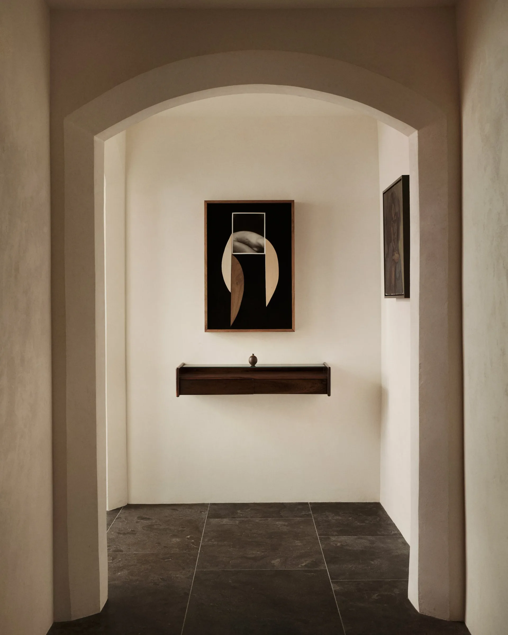 Minimalist gallery alcove with arched ceiling, dark flooring, and white walls displaying abstract artwork above a dark shelf by Ashe Leandro.